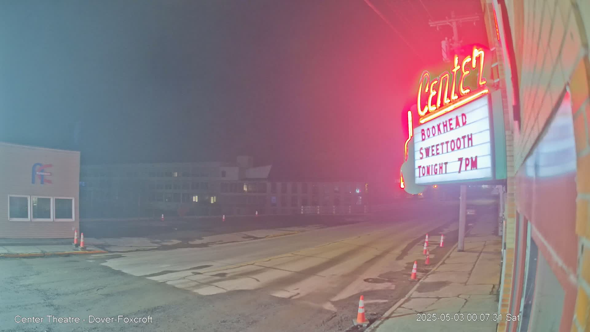 A fog-shrouded street at night shows a road, buildings, and a theater marquee with a neon sign.