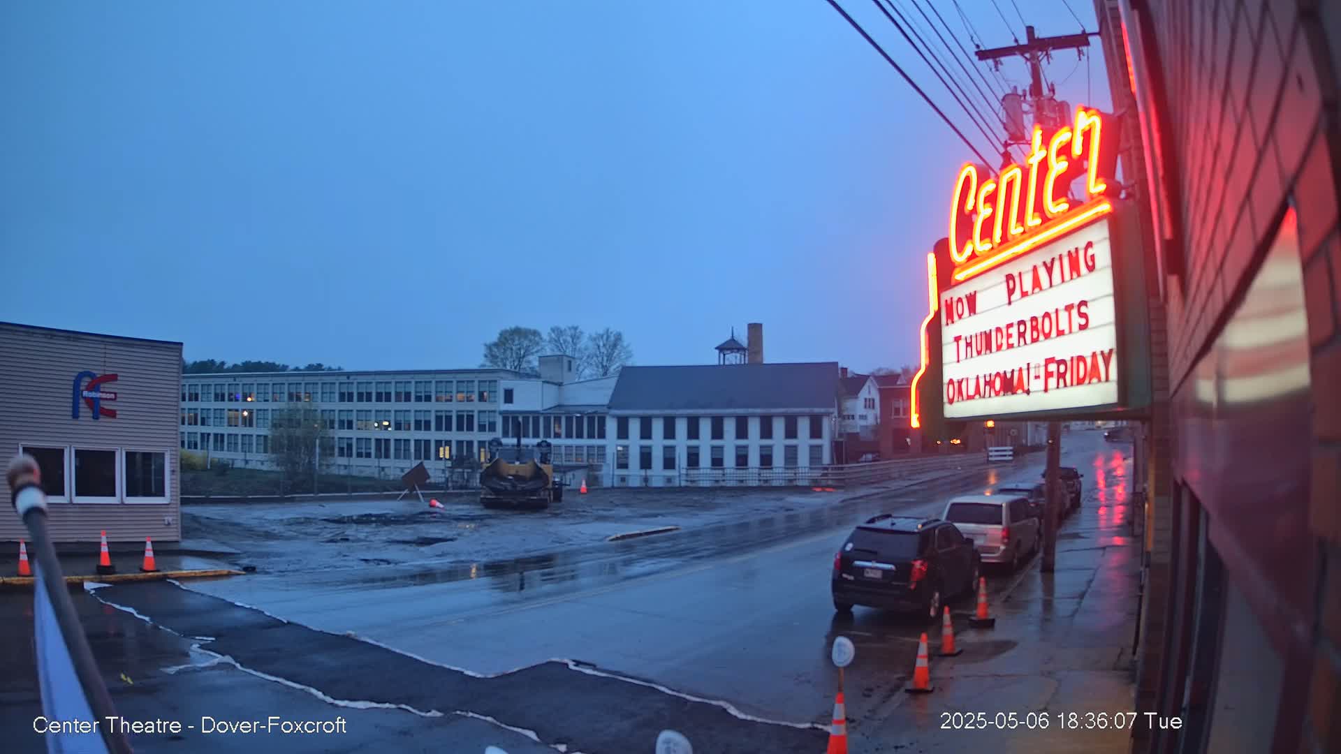 A wet street scene at dusk shows road construction near several buildings, including a movie theater with a marquee.
