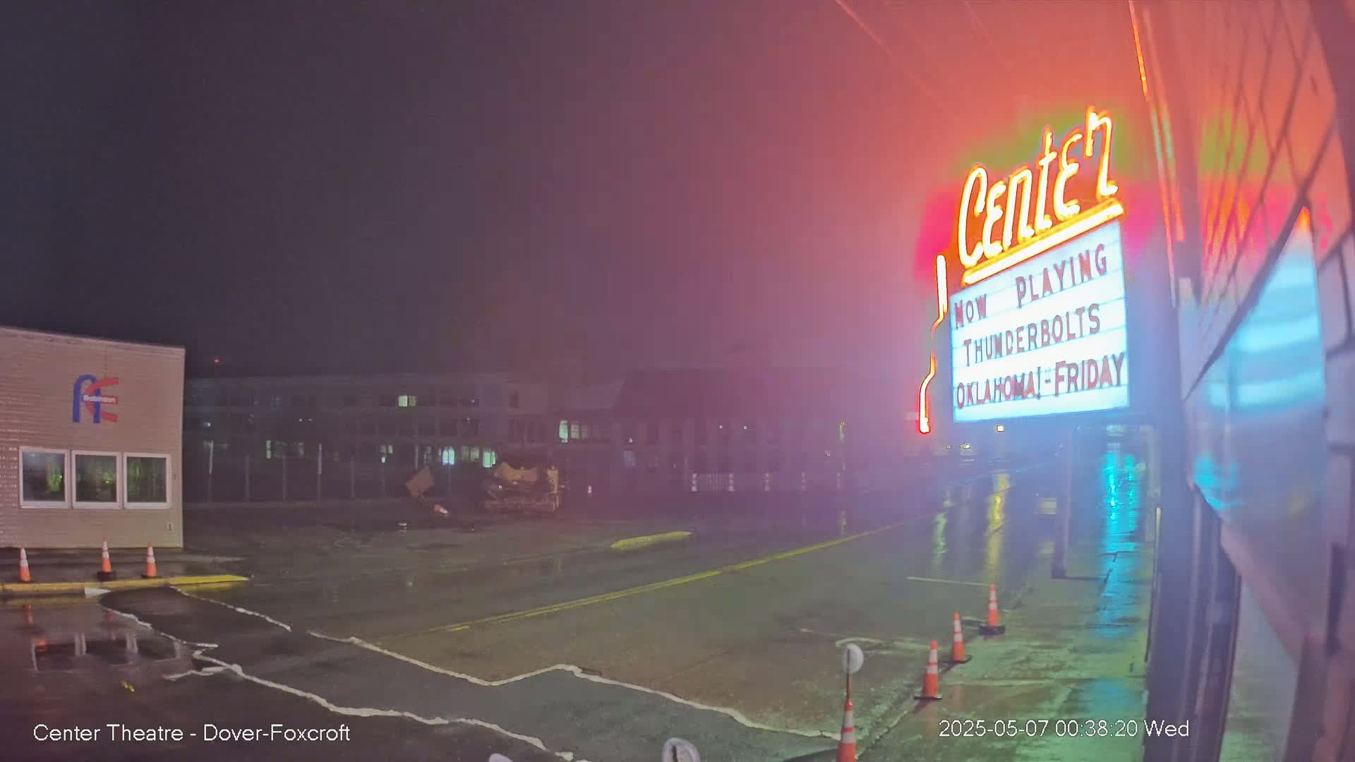 A rain-slicked parking lot at night features a movie theater marquee advertising "Thunderbolts" and "Oklahoma," with buildings and construction equipment visible in the background.