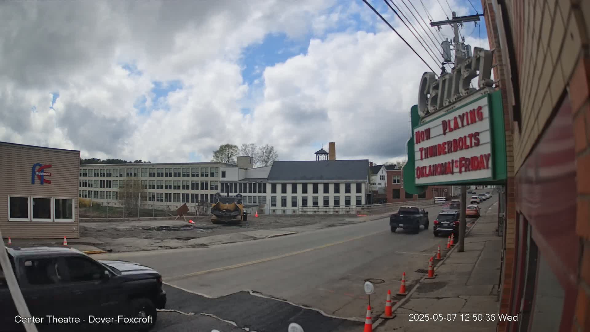 Under a partly cloudy sky, a street scene shows construction work in progress near a large school building and several other structures, with vehicles parked and driving on a road.