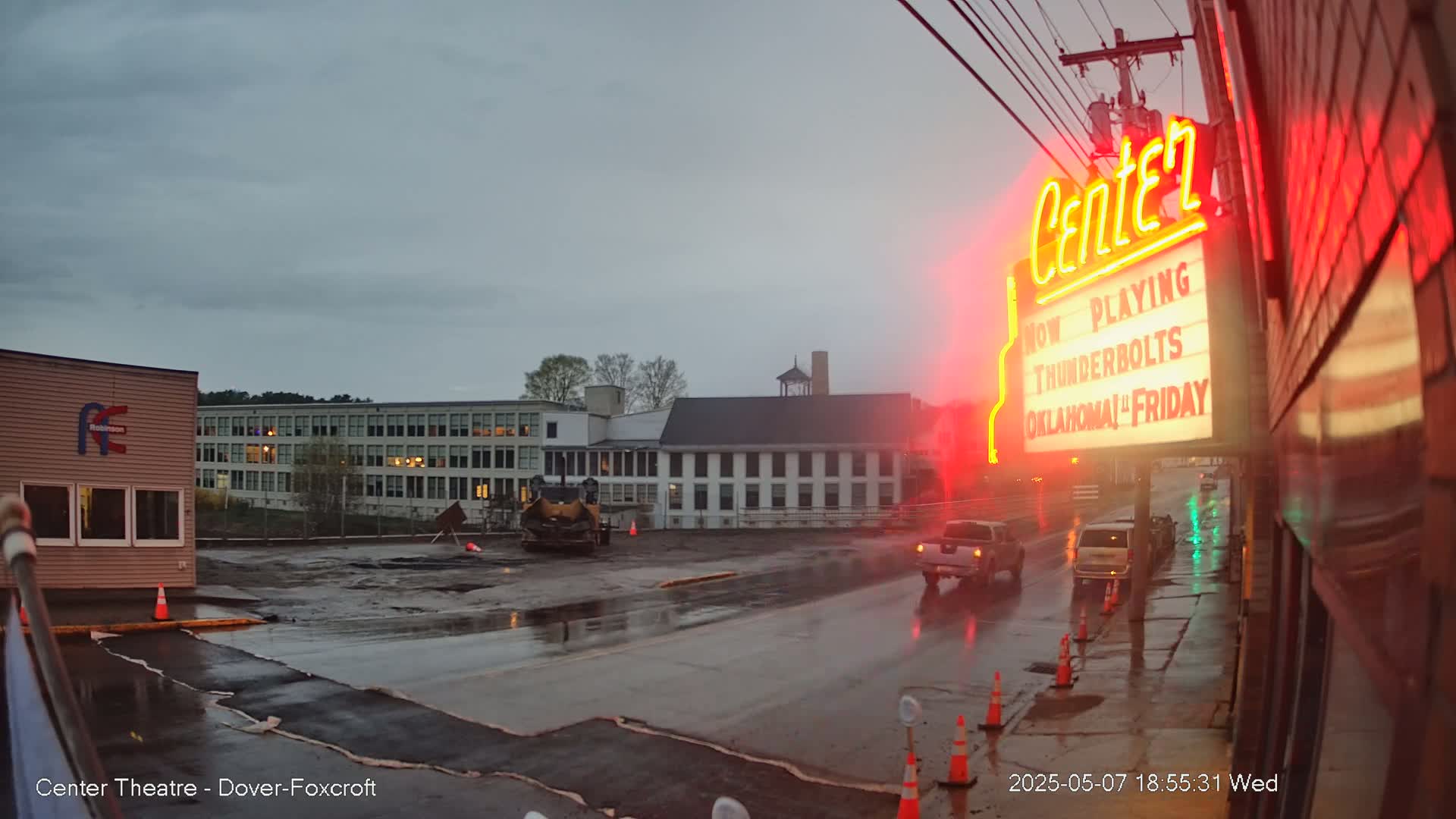 A wet street scene under an overcast sky shows road construction near a large building and a movie theater with a neon sign.