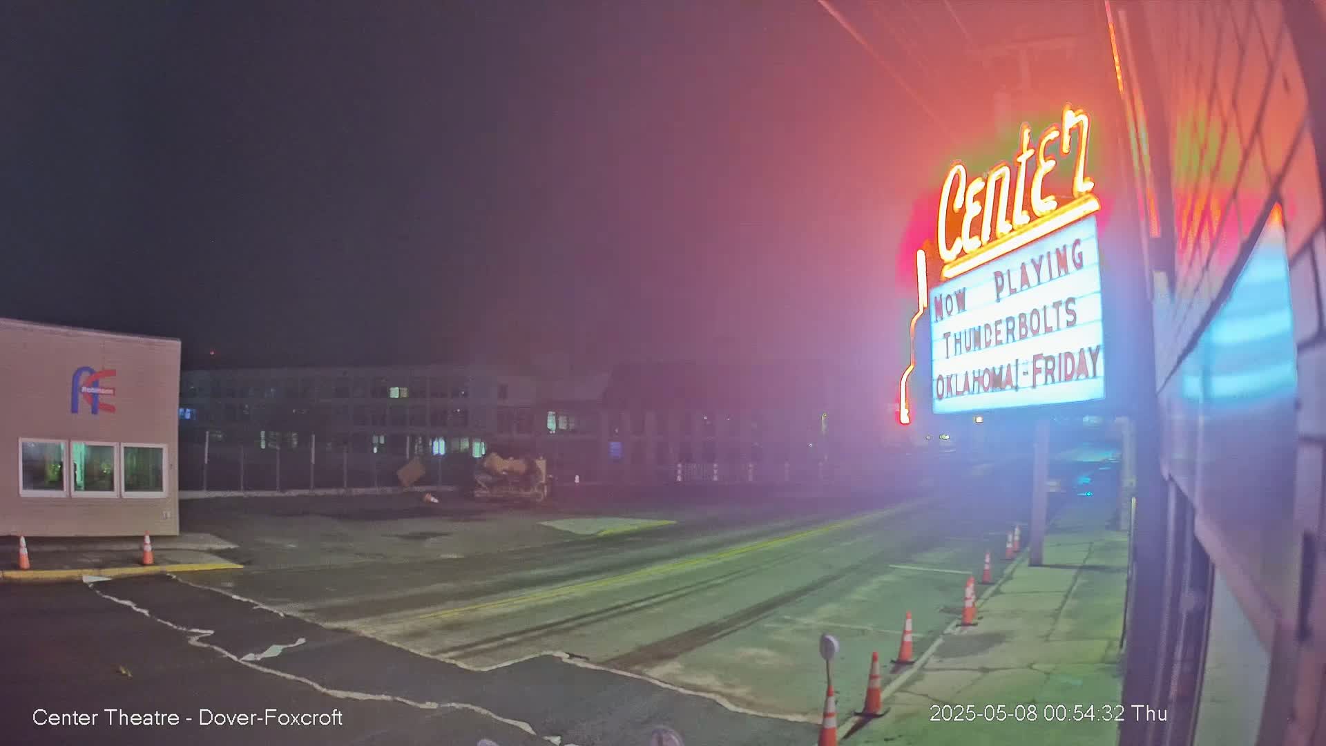 A nighttime, foggy view of a mostly empty parking lot in front of a movie theater marquee advertising "Thunderbolts" and "Oklahoma."