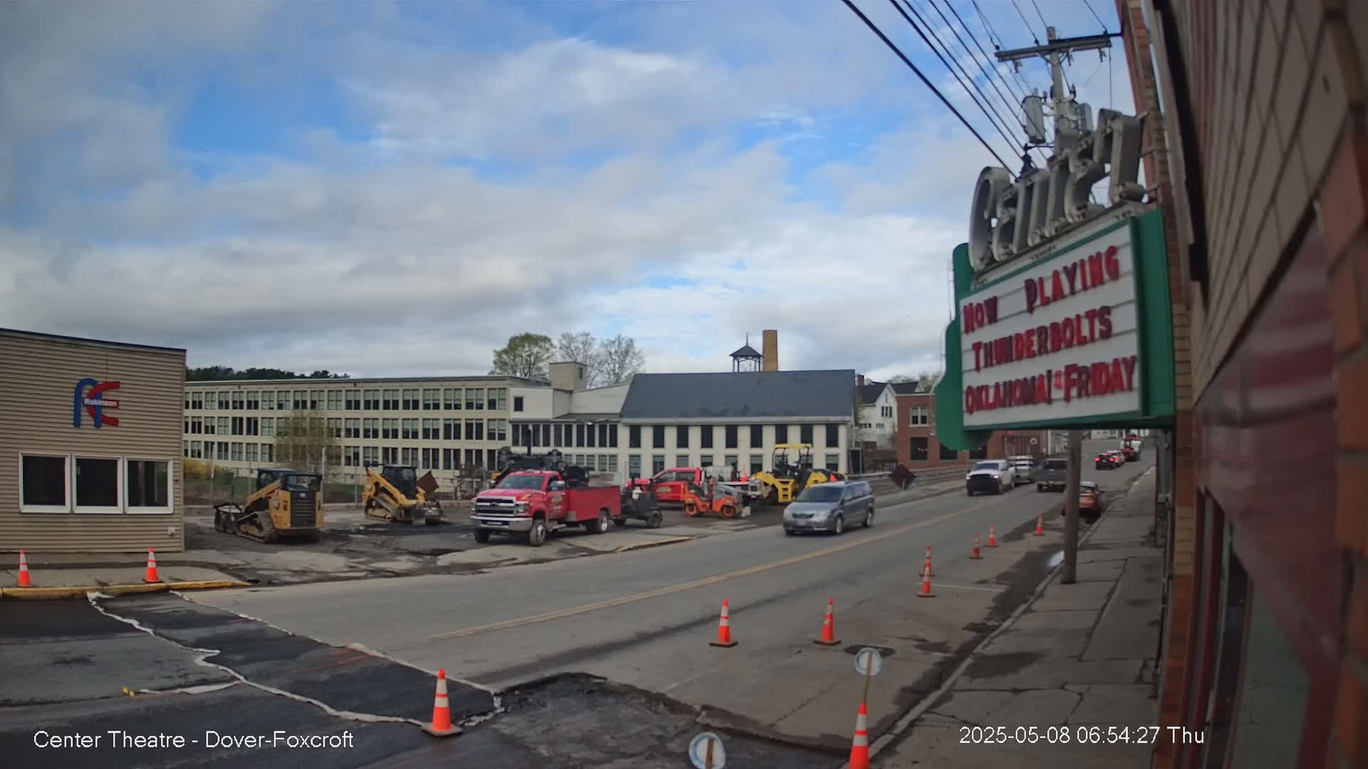 A street scene under partly cloudy skies shows construction workers and equipment near a large building and a movie theater.