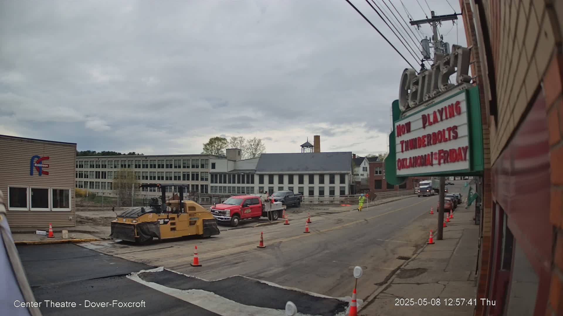 A street scene under an overcast sky shows road construction with a road roller and other vehicles, and buildings in the background.