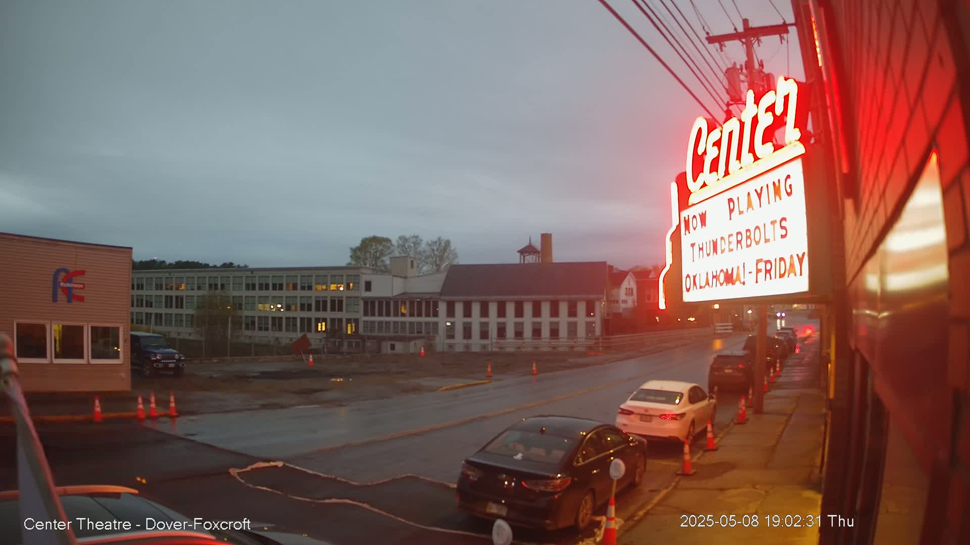 A street scene at dusk shows several cars parked along a curb near a theater marquee under a cloudy sky.