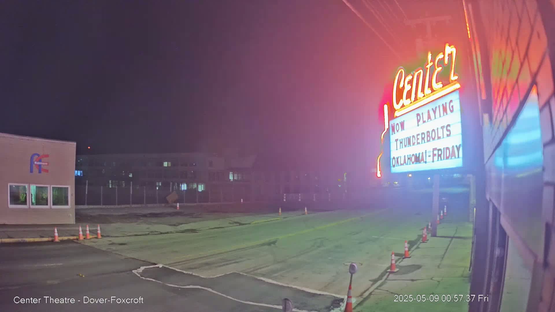 A dimly lit, empty street at night with a movie theater marquee showing "Thunderbolts" and orange traffic cones is visible in hazy conditions.