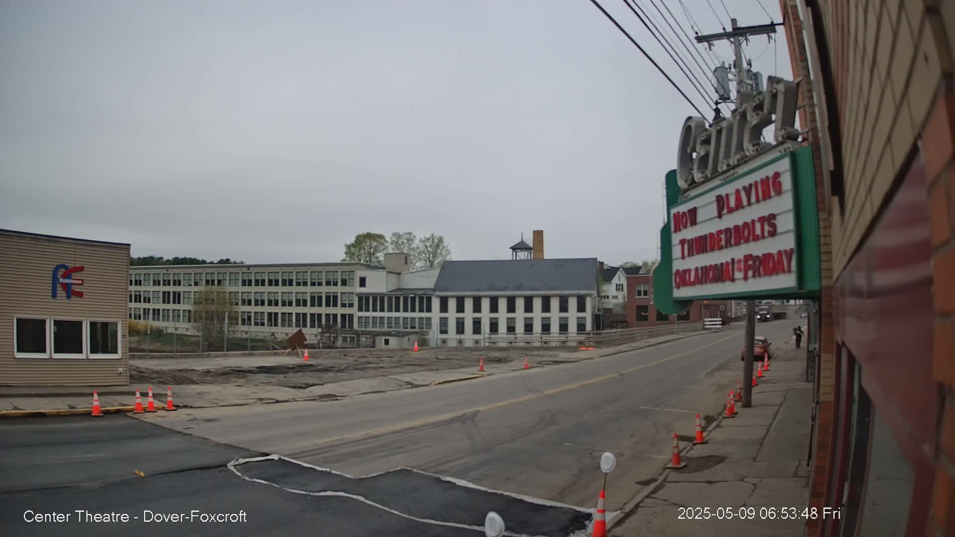 A mostly cloudy day reveals a street scene with construction in progress, featuring buildings of varying sizes and styles, including a large multi-story structure and a smaller one that appears to be a theater.