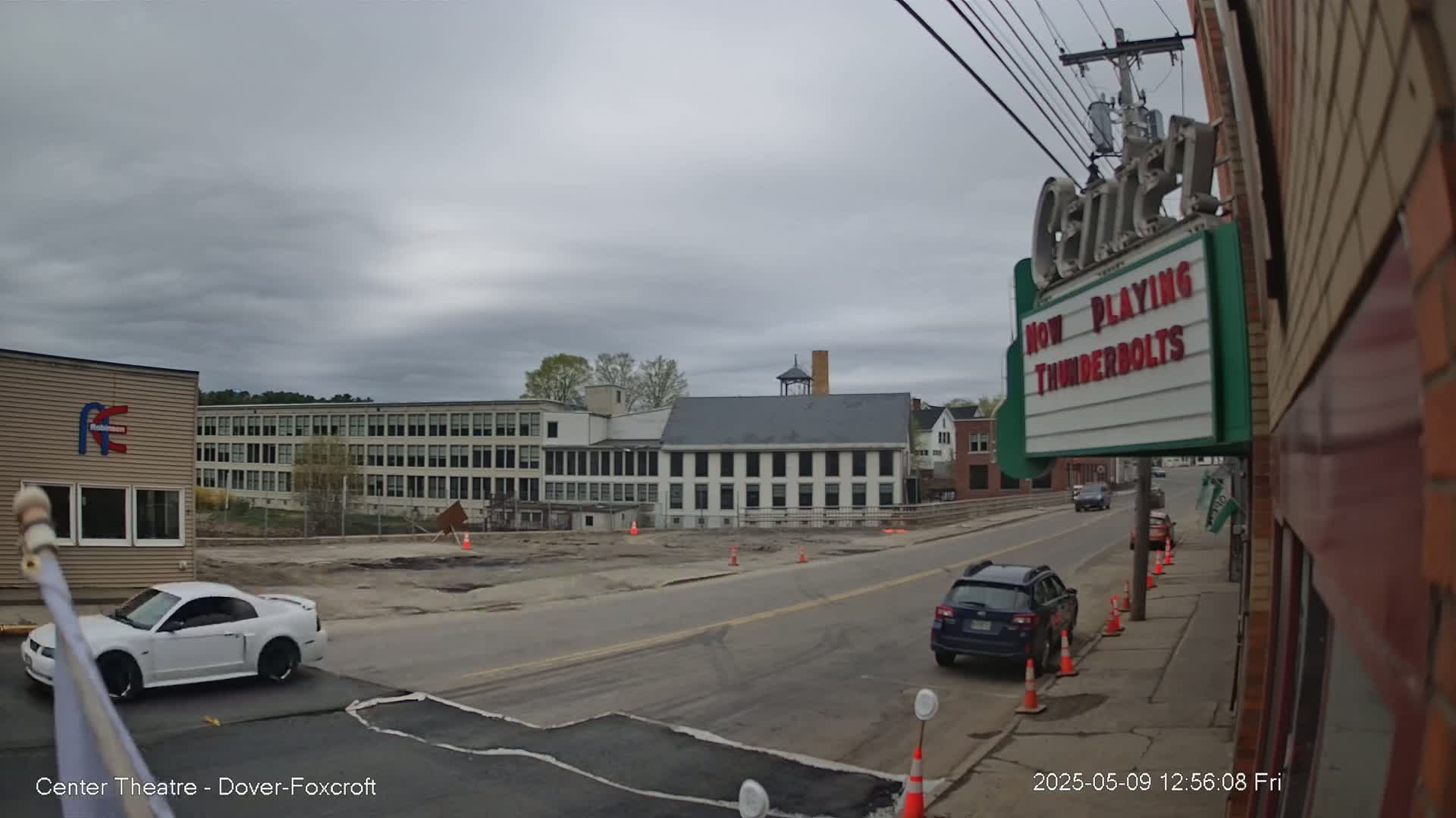 A street scene on an overcast day shows a white sports car parked near a building, with other cars driving on a road that passes in front of several older buildings, one of which is a theater with a marquee.