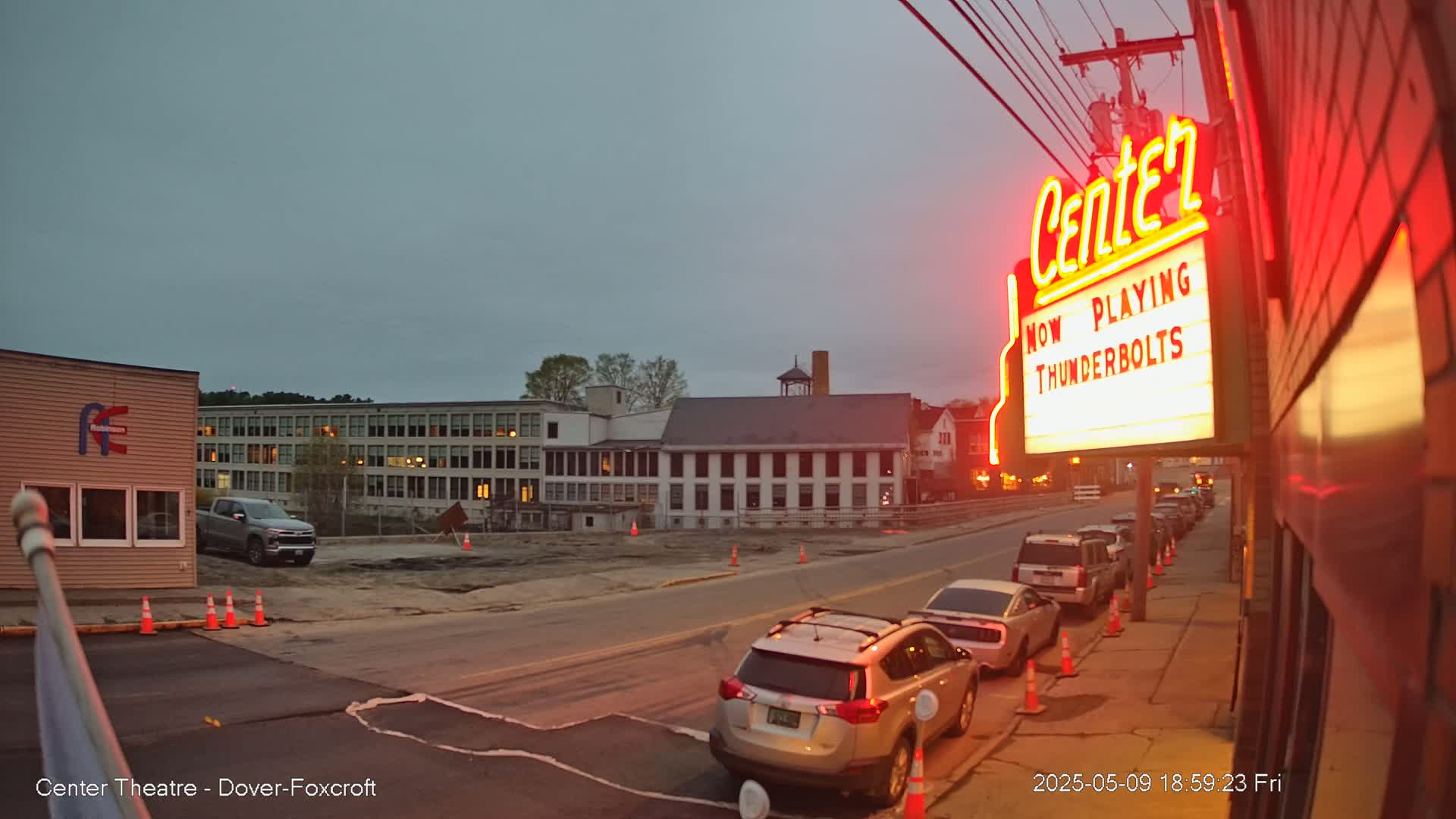 A movie theater marquee, illuminated in red, advertises "Thunderbolts," and several cars are parked along a street under a twilight sky.
