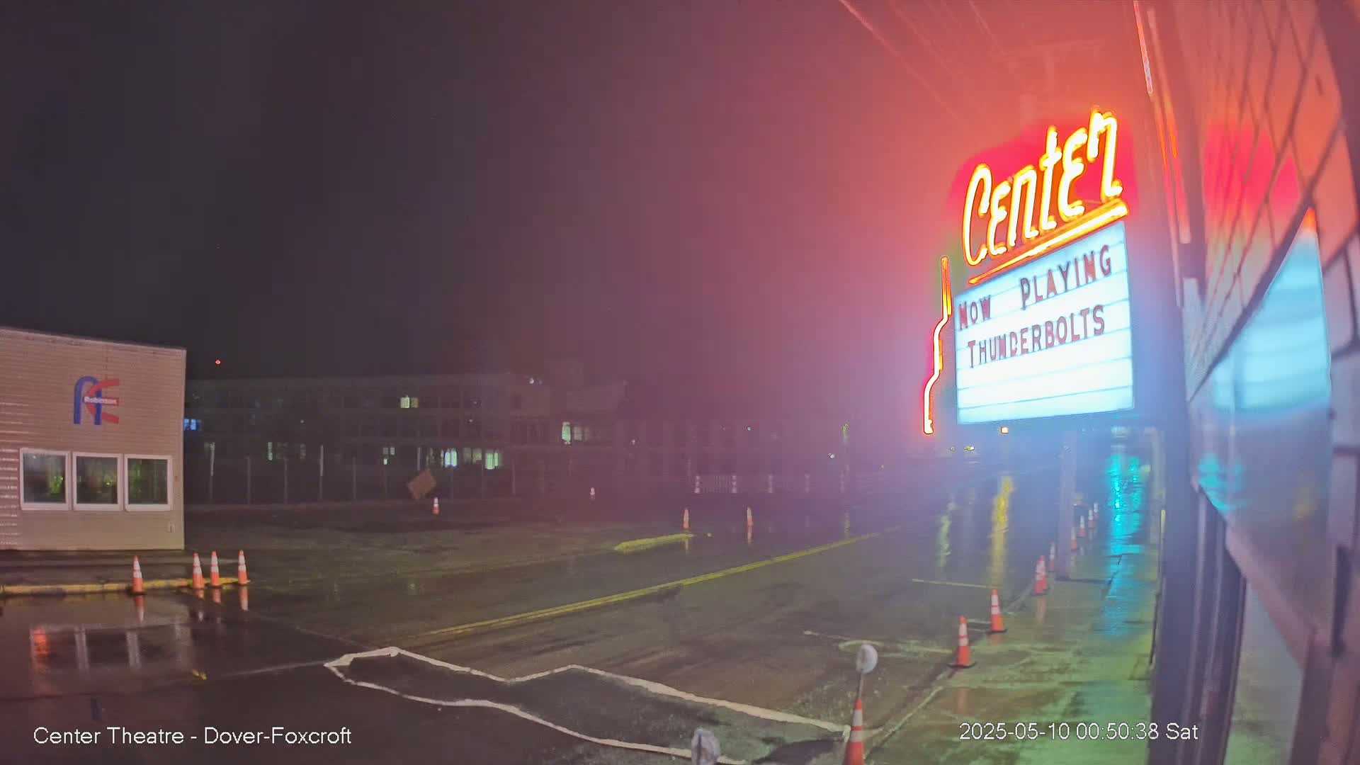 A wet, nighttime street scene shows a movie theater marquee advertising "Thunderbolts," with orange traffic cones lining the wet pavement in front of the building.