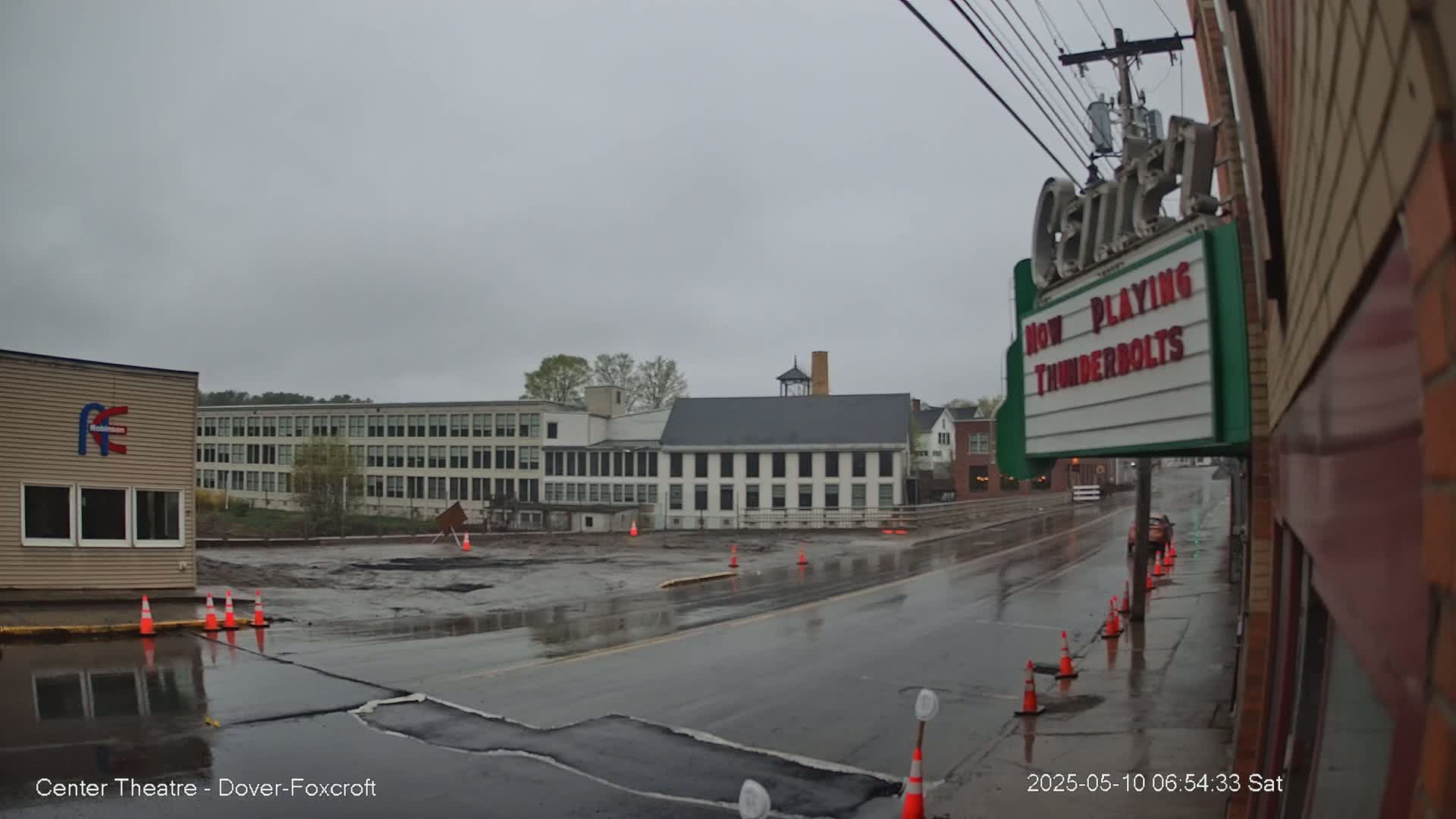 A wet street scene on an overcast day shows a theater marquee, several buildings, and construction cones lining a roadway under repair.