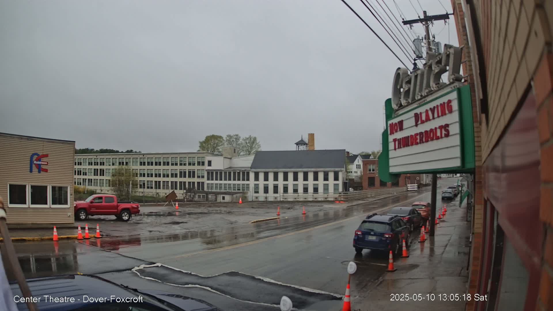 A wet street scene shows several cars parked along the curb, a red pickup truck in a parking lot, and a large multi-story building across the street under a gray, overcast sky.