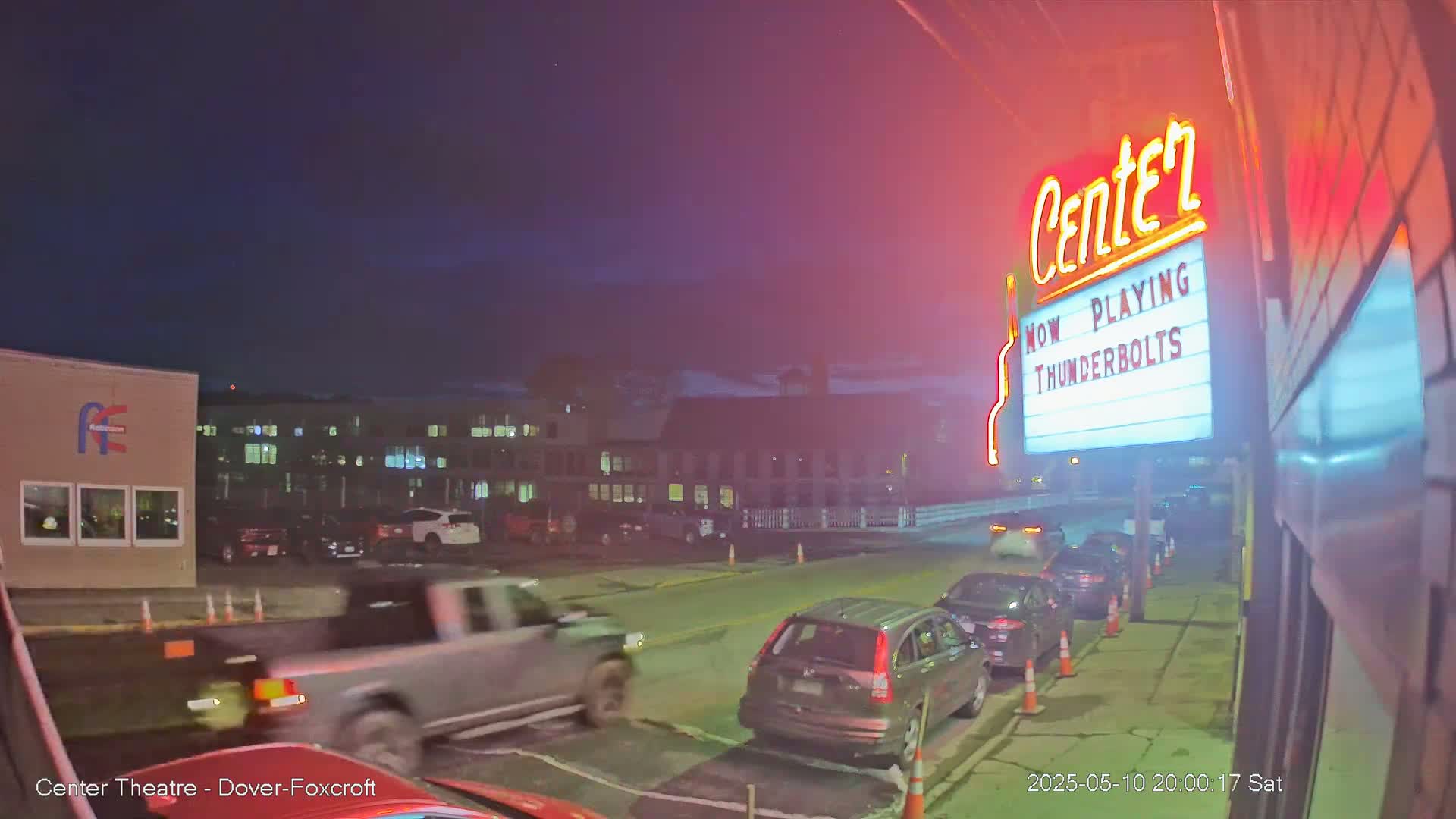 At night, several cars are parked in a line in front of a movie theater marquee under a dark, mostly clear sky.