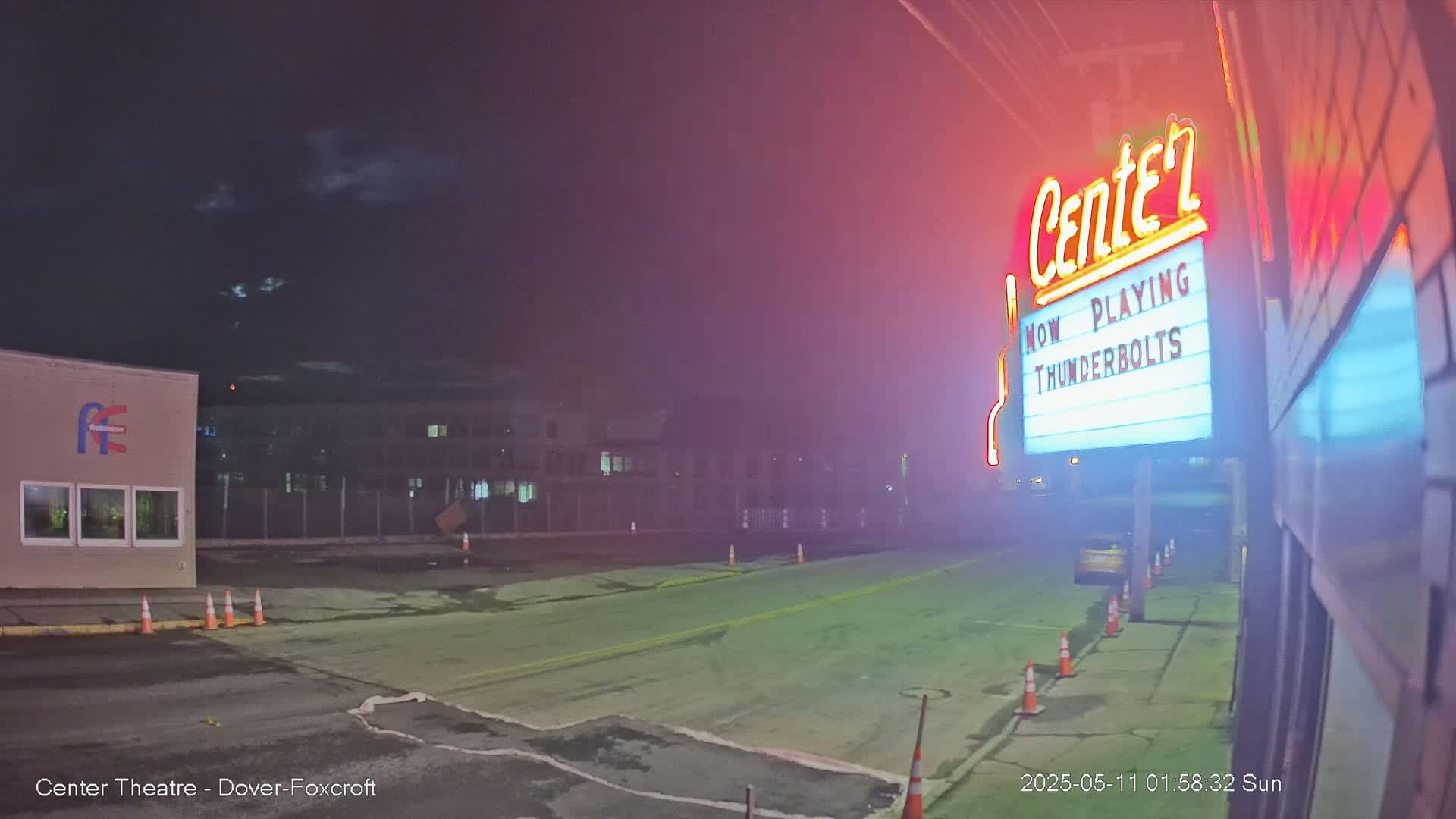 A nighttime view of a mostly empty street in front of a movie theater marquee displaying "Thunderbolts," under a partly cloudy sky.