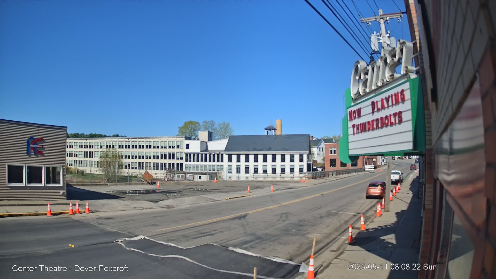 A sunny day reveals a street scene with several buildings, including a large factory, a smaller brick building, and a theater marquee, with a few cars parked along the roadside and construction cones scattered about.