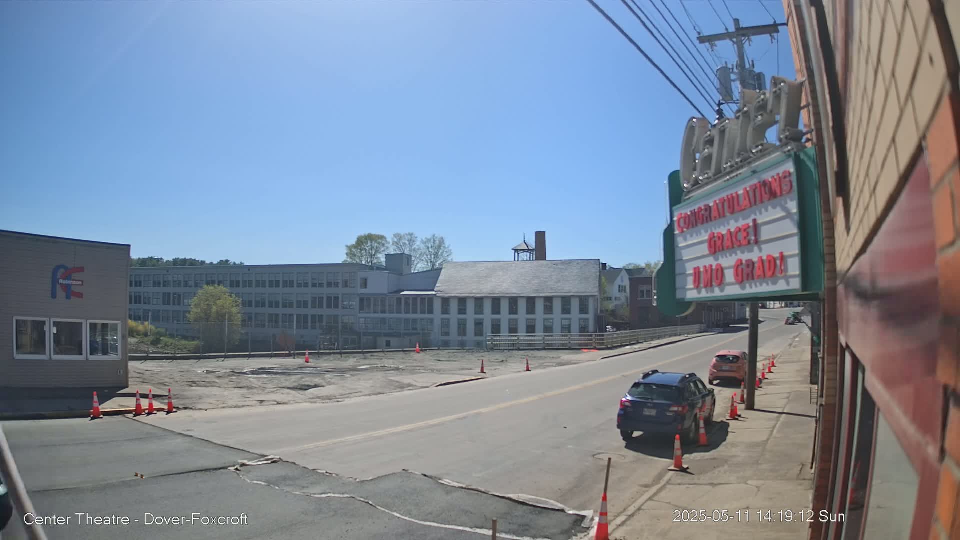 A sunny day reveals a street scene with parked cars, a large building under construction, and a marquee displaying a graduation message.