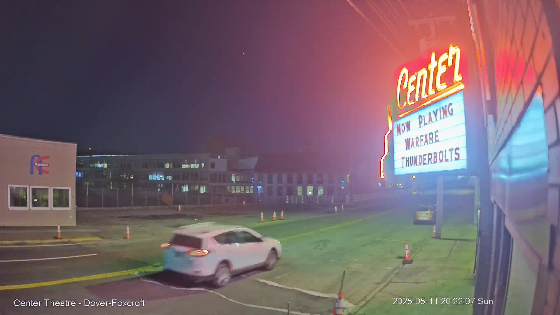 A white SUV drives past a movie theater marquee at night under a clear sky.