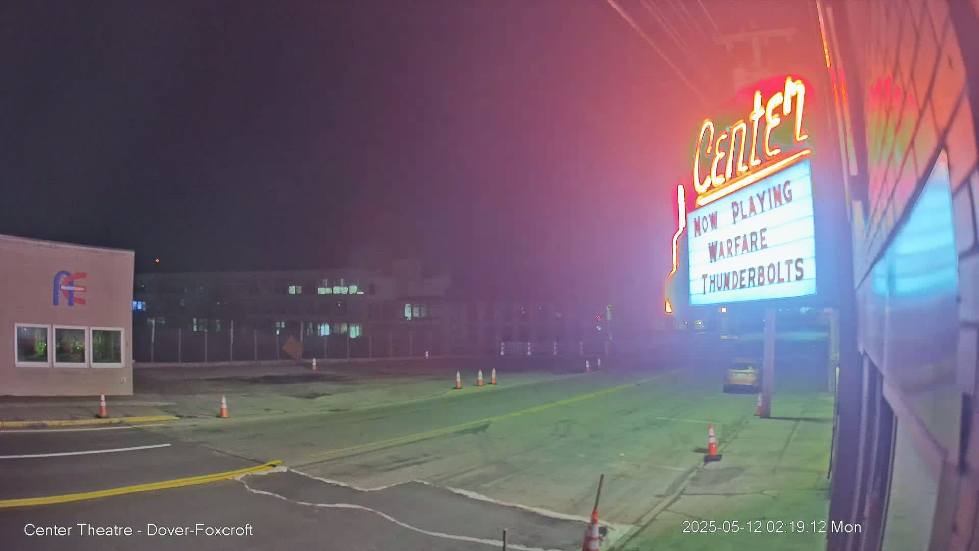 A nighttime, hazy view of a mostly empty street in front of a movie theater marquee advertising "Warfare Thunderbolts."