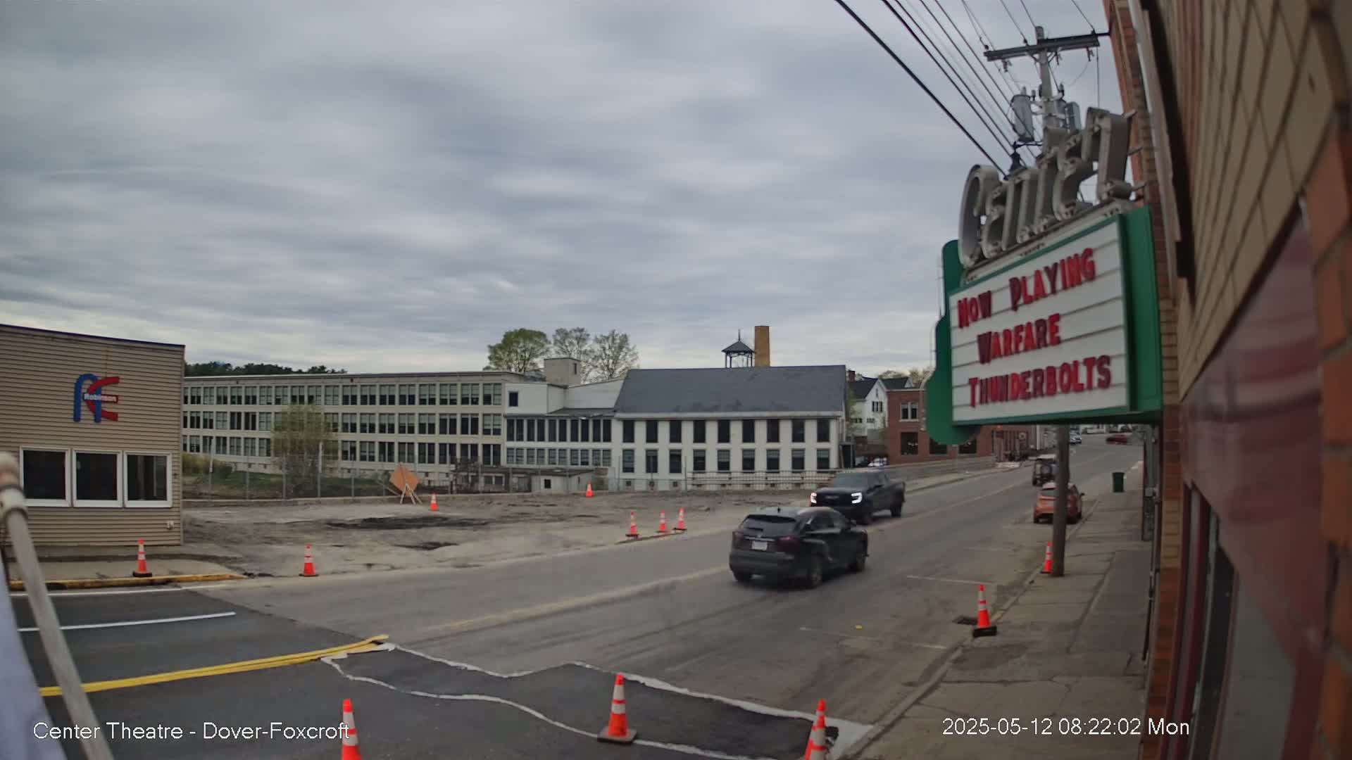 Under an overcast sky, two cars drive down a street past a partially cleared construction site, with a large building and a movie theater in the background.