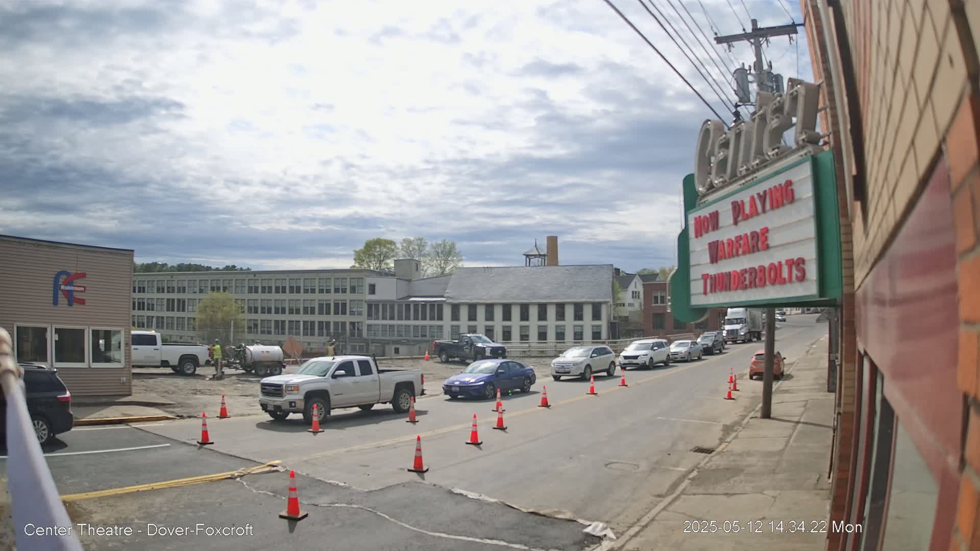 A street scene under a partly cloudy sky shows roadwork in progress near several buildings, including a large multi-story structure and a movie theater marquee.