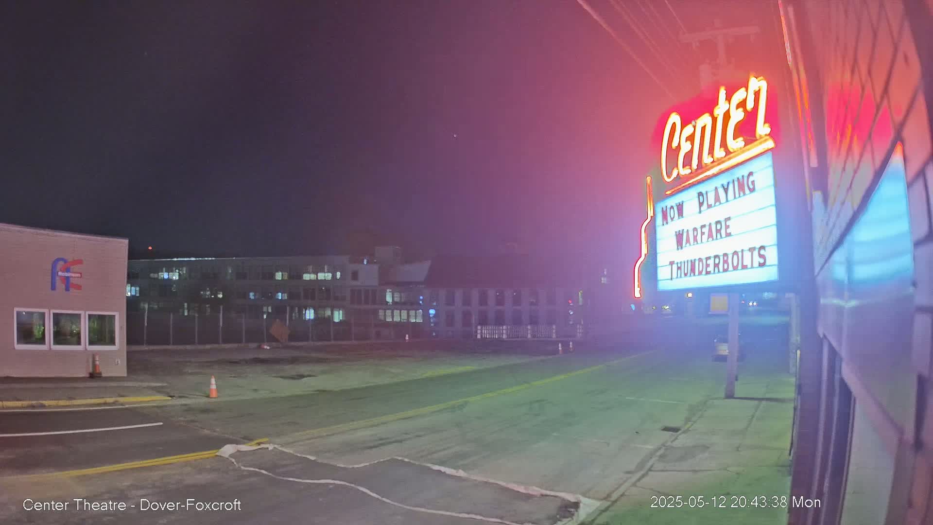 A nighttime view of a mostly empty street in front of a movie theater marquee advertising "Warfare Thunderbolts," with hazy conditions and buildings in the background.