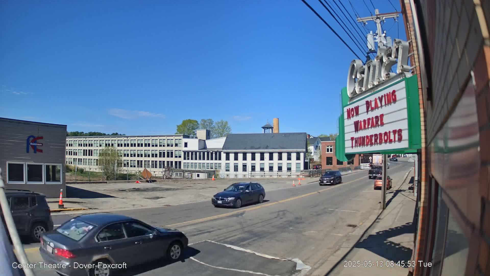 A sunny day shows a street scene with several cars, and buildings including an industrial-looking structure and a movie theater marquee.