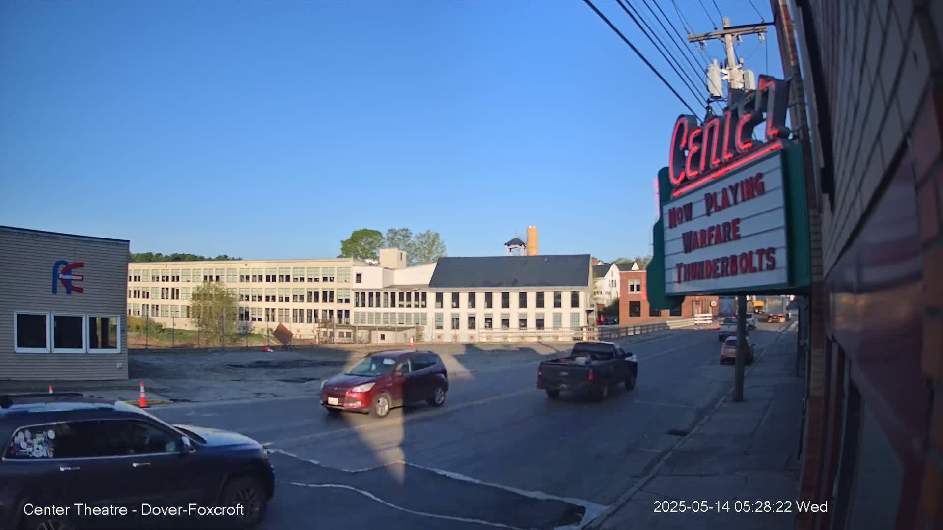 Under a clear blue sky, a street scene shows several cars driving past a large building and a movie theater marquee.