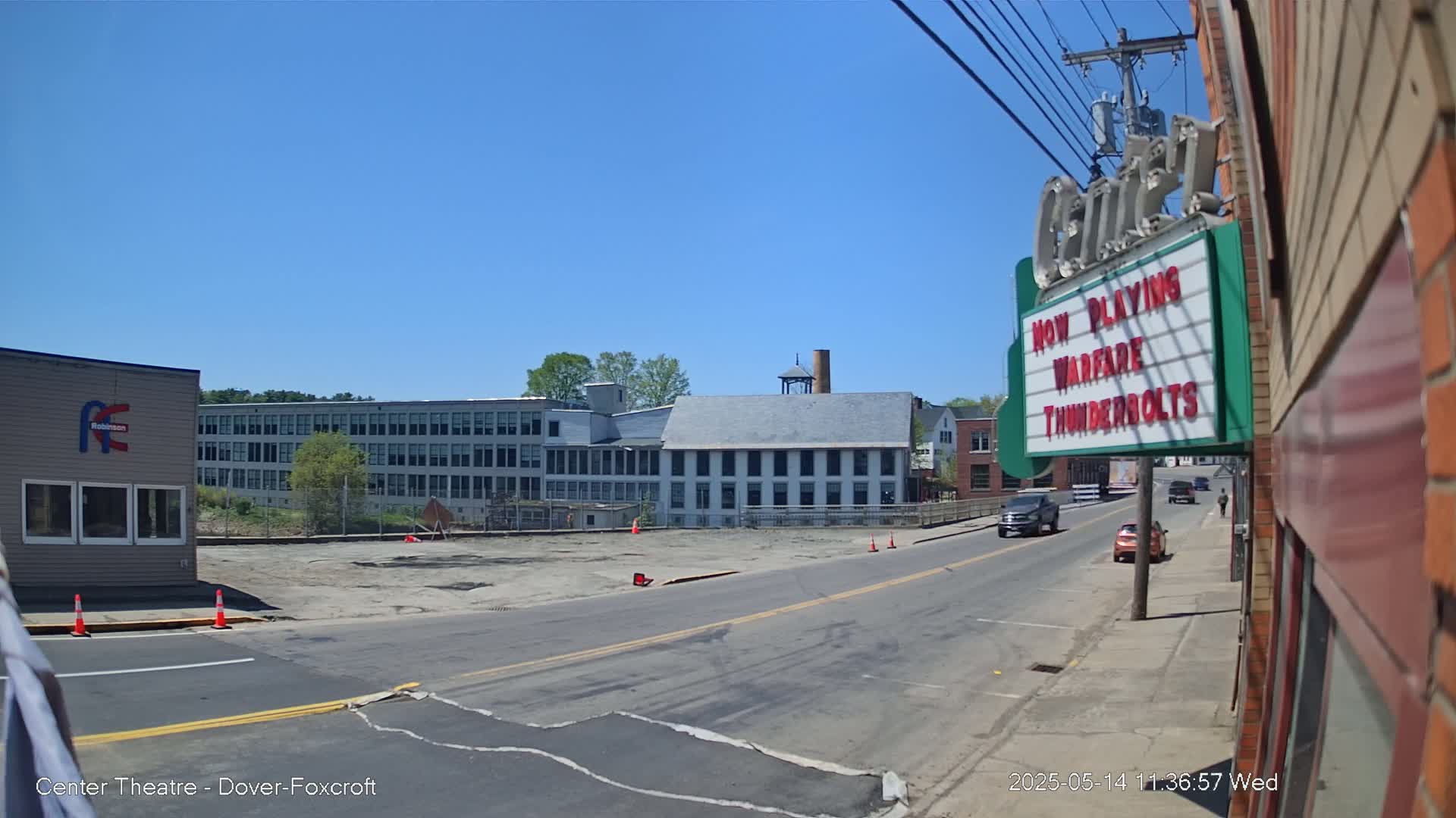 A sunny day reveals a street scene with a large empty lot, several industrial-style buildings, and a couple of cars driving on the road.
