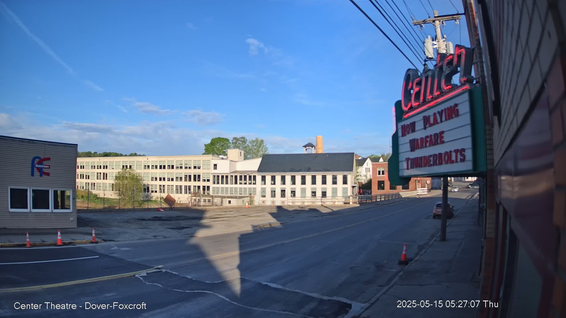 A mostly sunny street scene shows several buildings, including a large factory, under a clear blue sky.