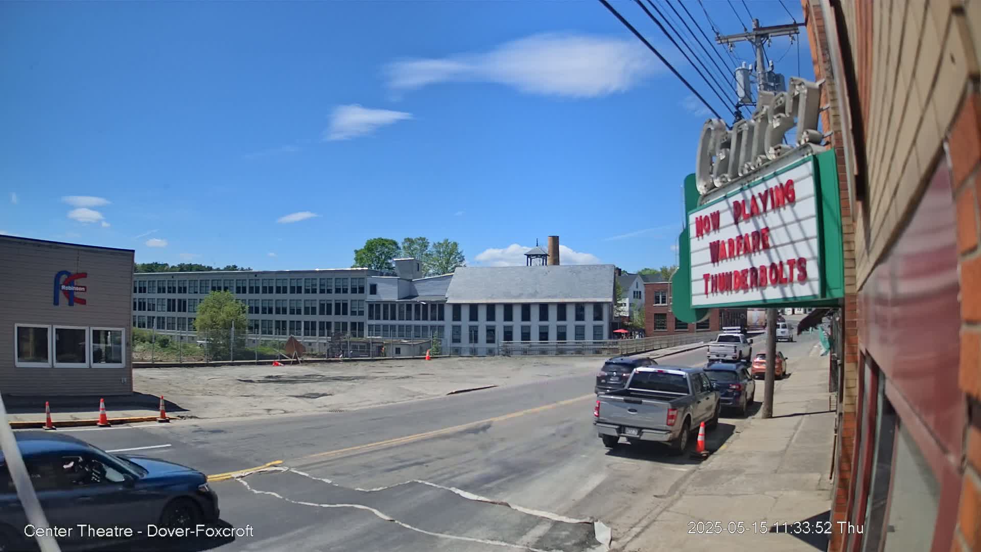 A sunny day reveals a street scene with several cars parked along the curb, and a large industrial building and smaller buildings across the street.