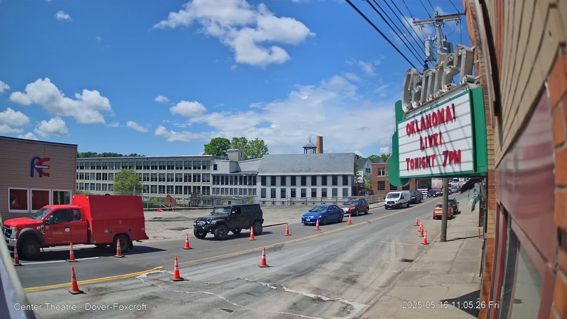 Under a mostly sunny sky, a street scene shows several vehicles, including a red utility truck, stopped or slowing near construction cones in front of a large brick building.