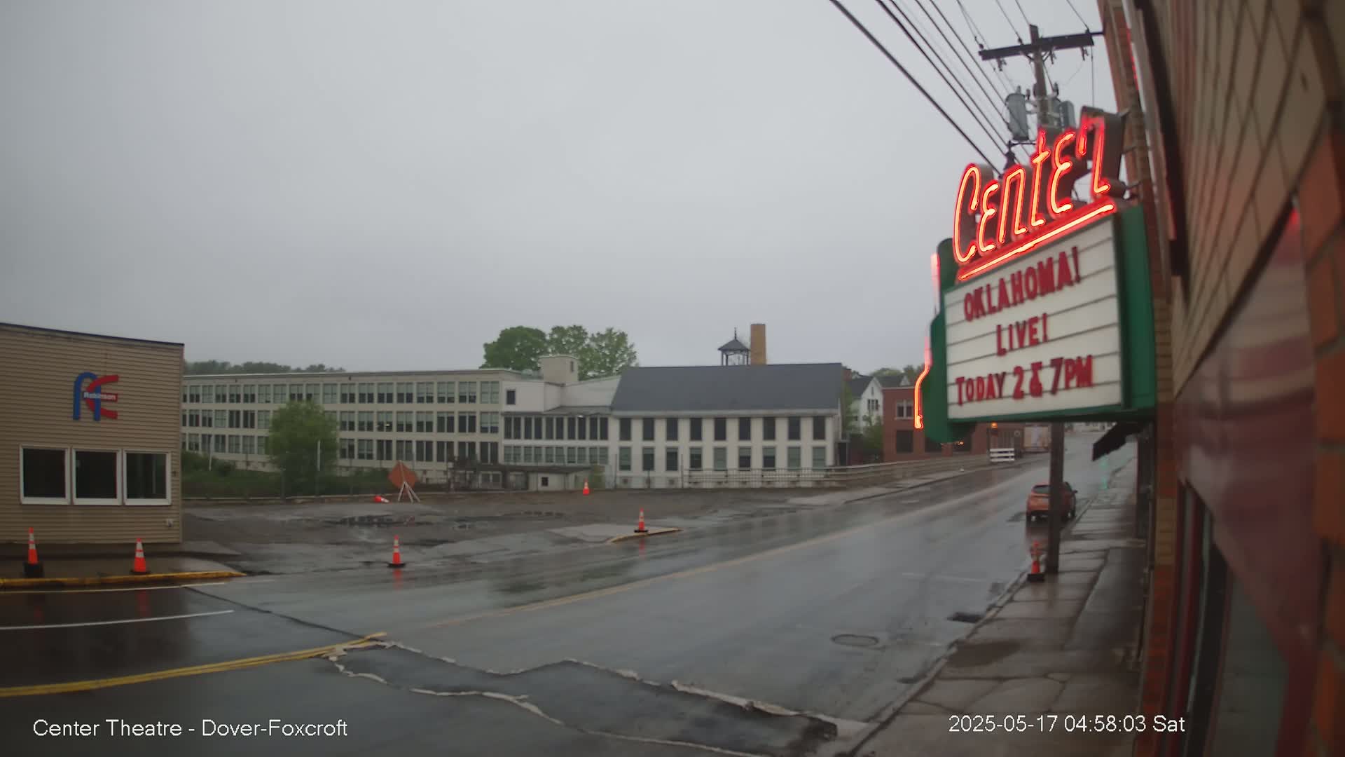 A rain-slicked street scene shows several buildings under an overcast sky, with a theater marquee advertising a live show.