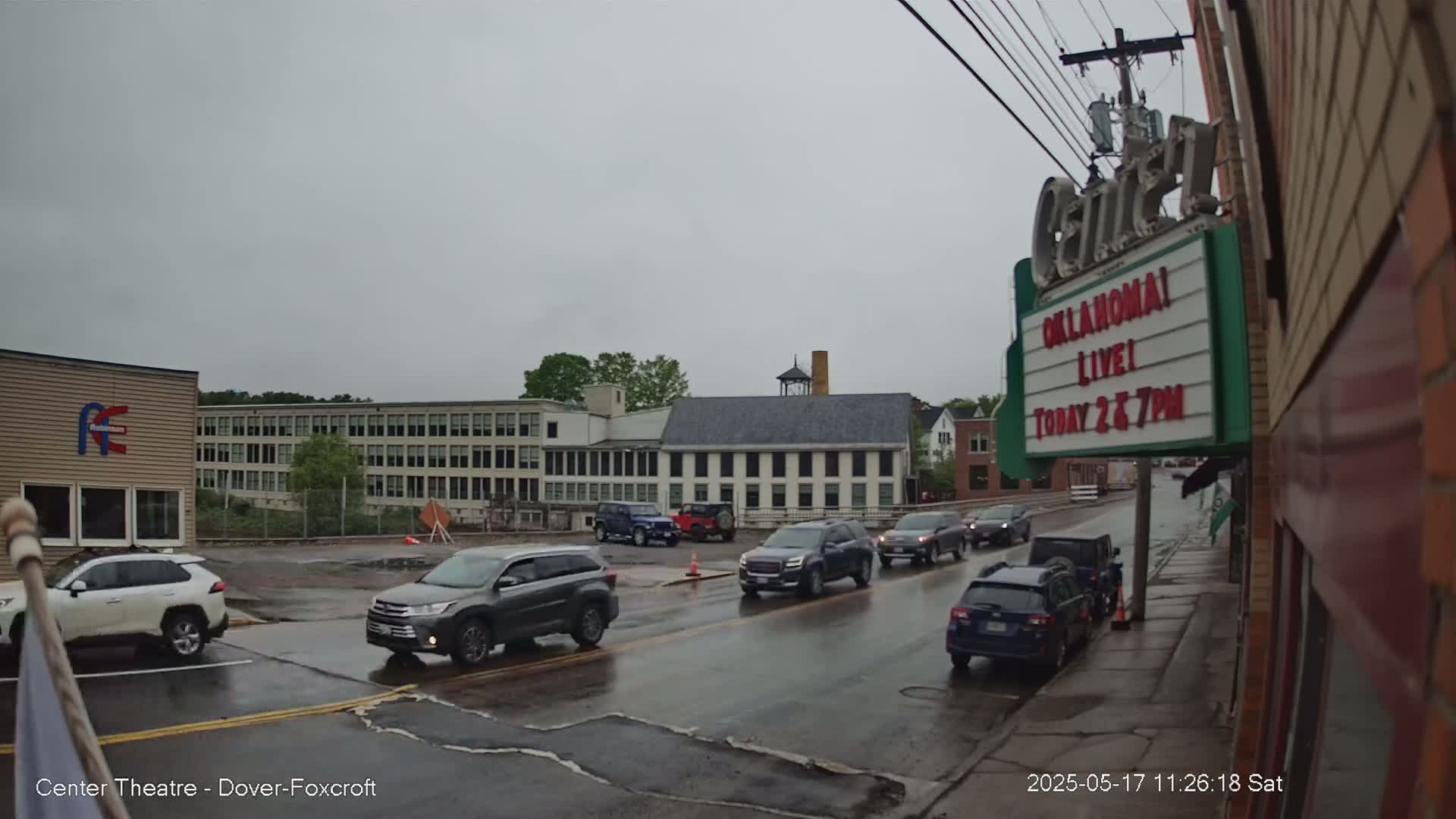 Several cars drive on a rain-slicked street past multi-story buildings under a cloudy sky.