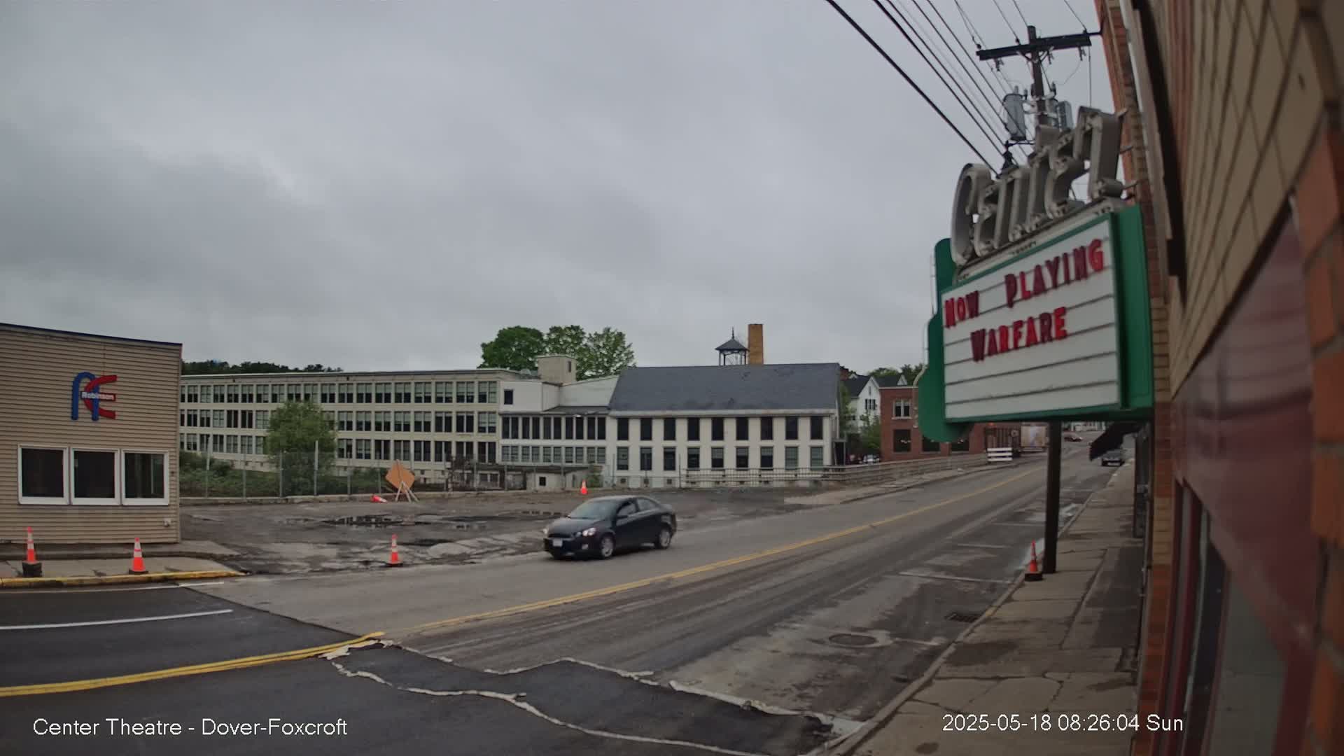 A dark-colored car drives down a street past a movie theater marquee advertising "Warfare," under an overcast sky, with construction visible in the foreground and older buildings in the background.