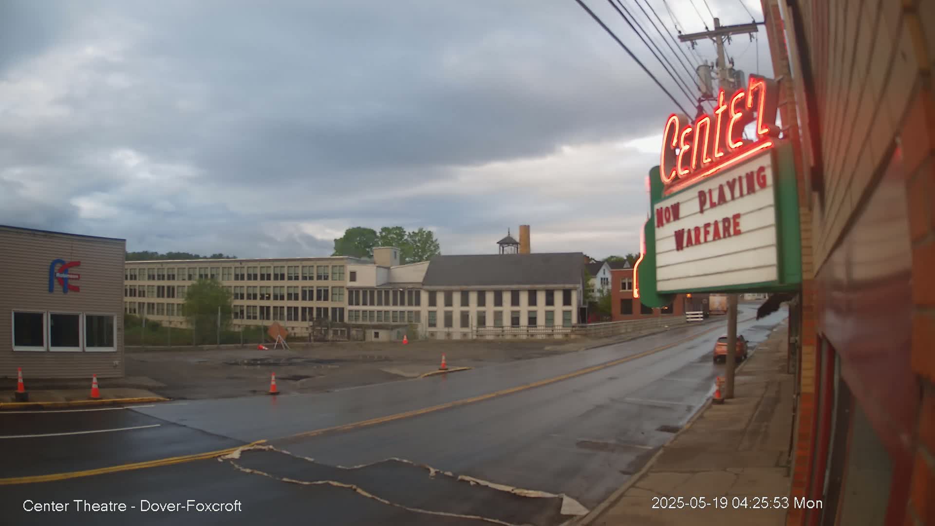 A wet street scene under a cloudy sky shows a movie theater marquee advertising "Warfare," industrial buildings, and a car parked on the roadside.