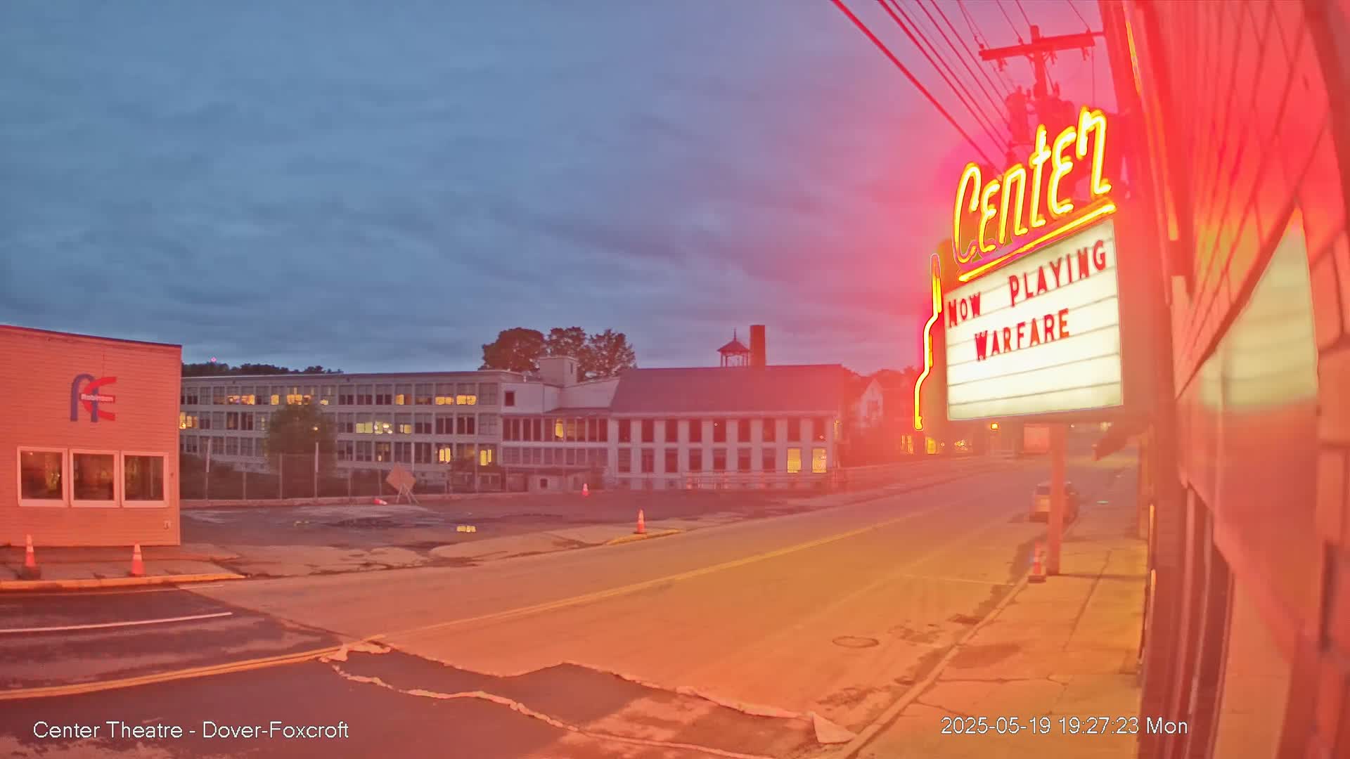 Under a twilight sky, a street scene shows a movie theater marquee advertising "Warfare," with a multi-story building and other structures visible in the background.