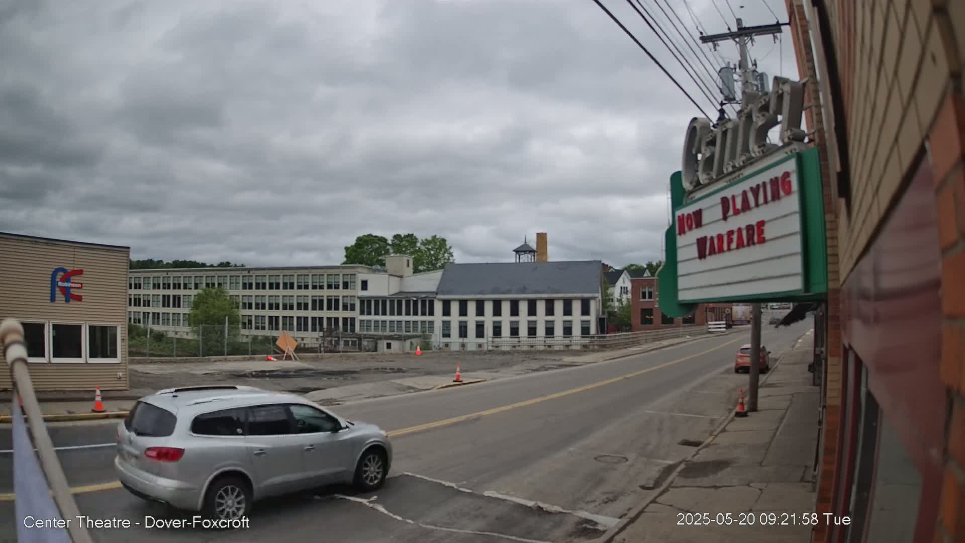 A silver SUV drives past a movie theater marquee on a street under a cloudy sky, with industrial buildings visible in the background.