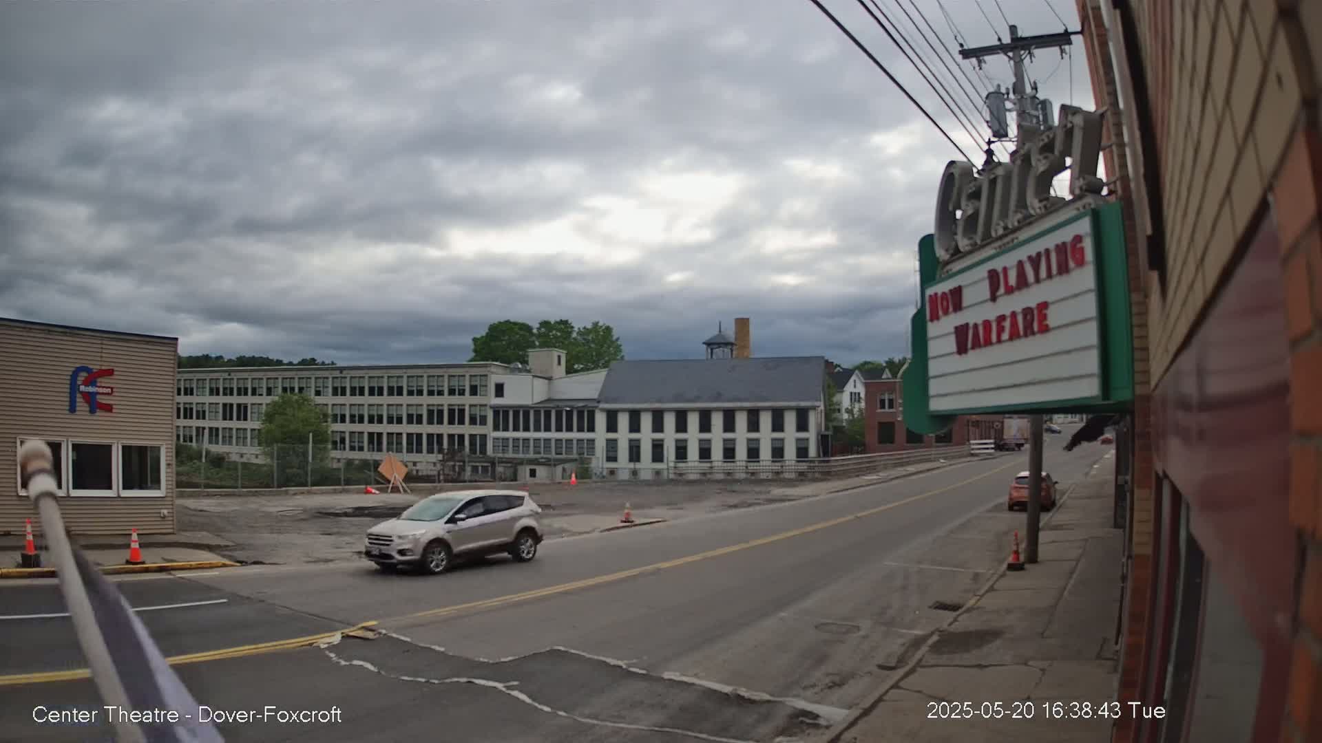 A silver SUV drives past a street scene under a cloudy sky, with large industrial buildings and a movie theater marquee in the background.