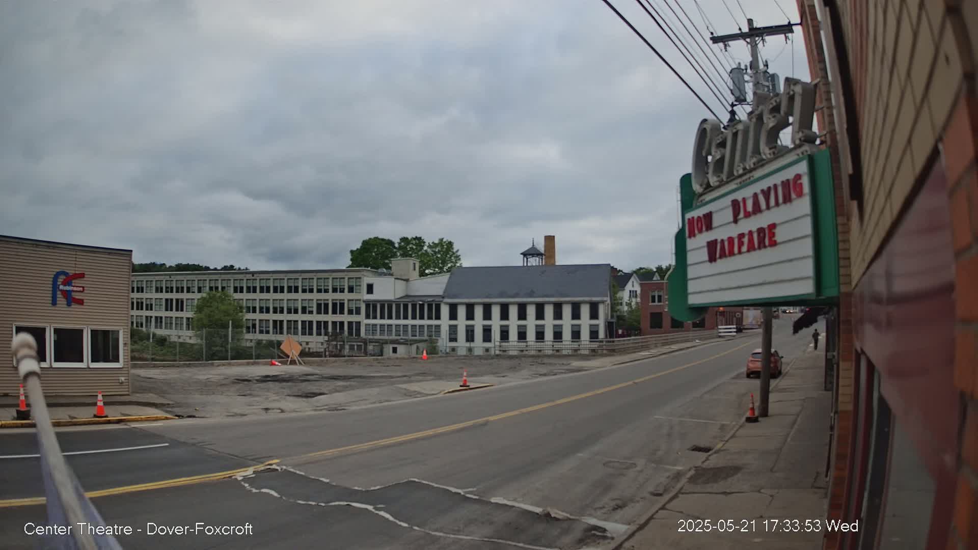 A street scene under a cloudy sky shows a road leading to several multi-story buildings, one of which is a movie theater displaying "Warfare" as the current film.