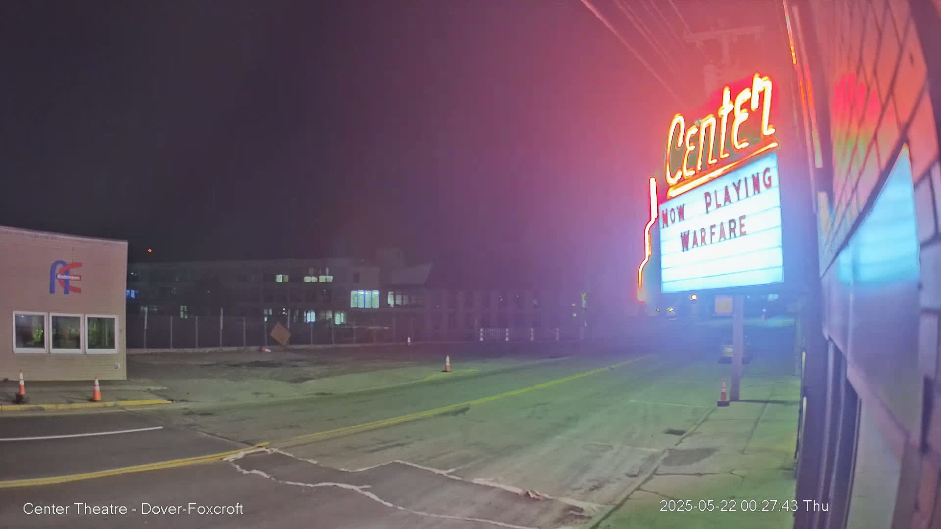 A nighttime street scene shows a mostly empty parking lot in front of a movie theater marquee advertising "Warfare," with a building visible in the background under a dark sky.