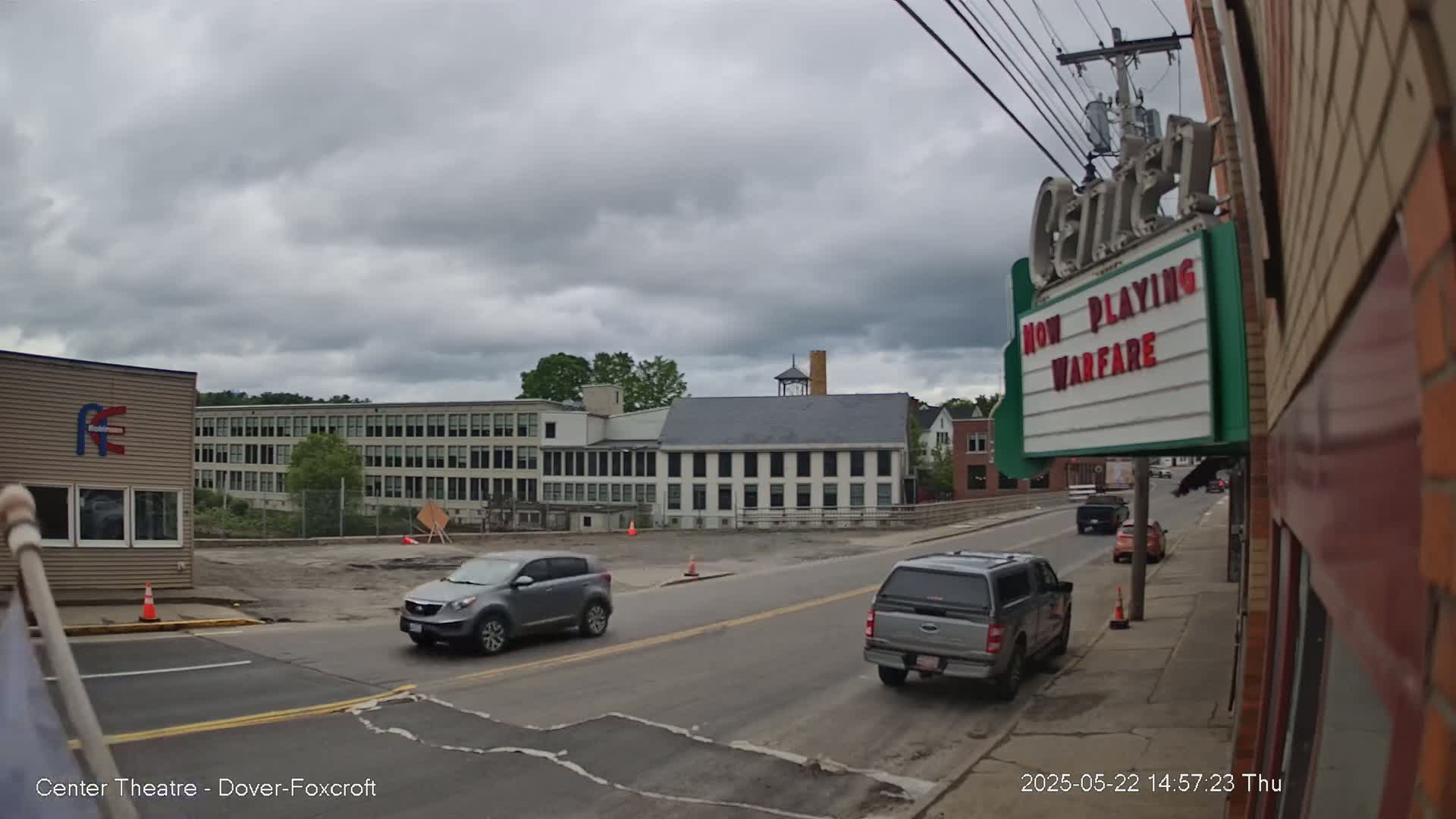 A street scene under a cloudy sky shows several vehicles driving on a road, passing in front of a large building and a movie theater marquee advertising "Warfare."