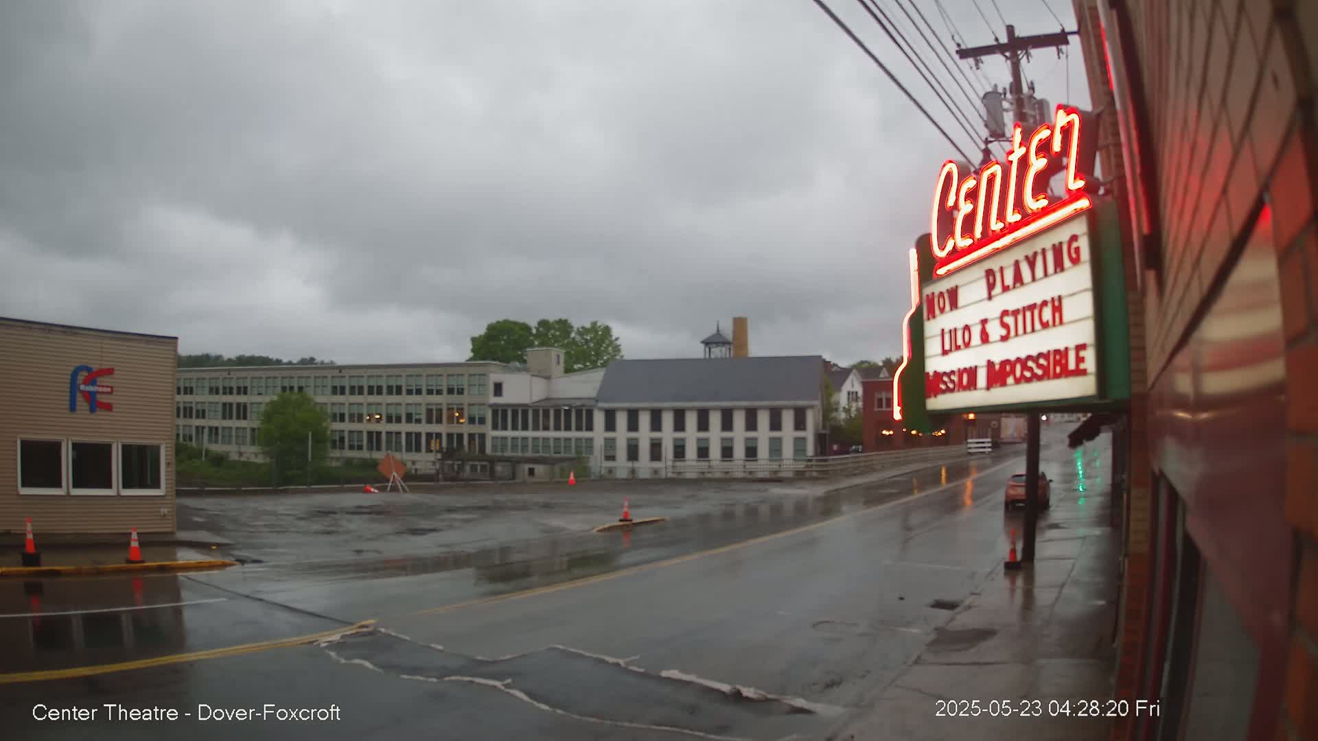 A wet street scene under an overcast sky shows a movie theater marquee advertising "Lilo & Stitch" and "Mission Impossible," with several buildings visible in the background.