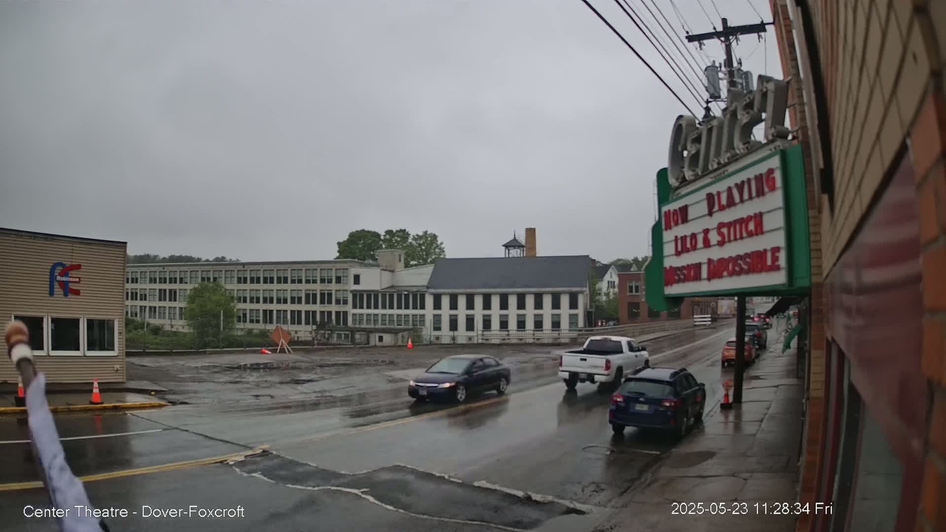 A rainy street scene shows several cars driving past a movie theater marquee and older buildings.