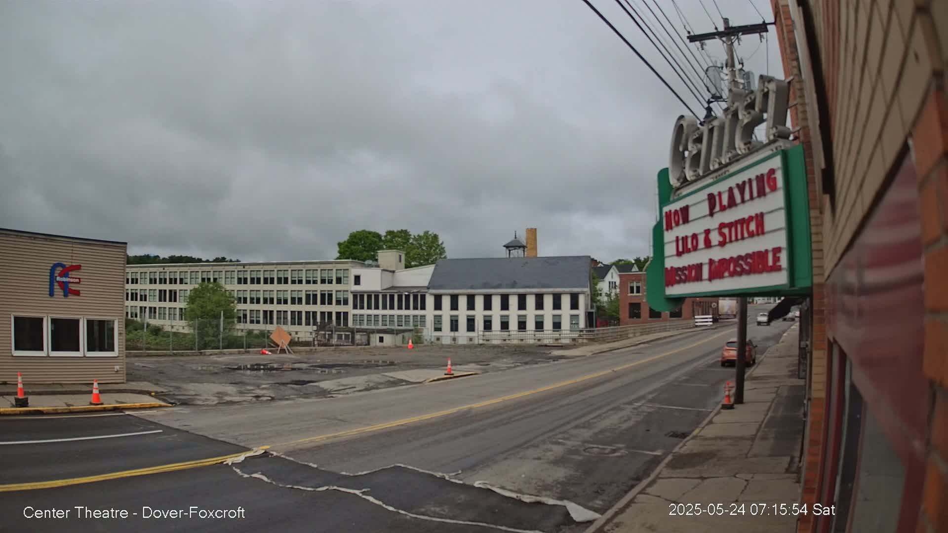 A street scene on an overcast day shows a mostly empty road in front of a movie theater and several multi-story buildings.