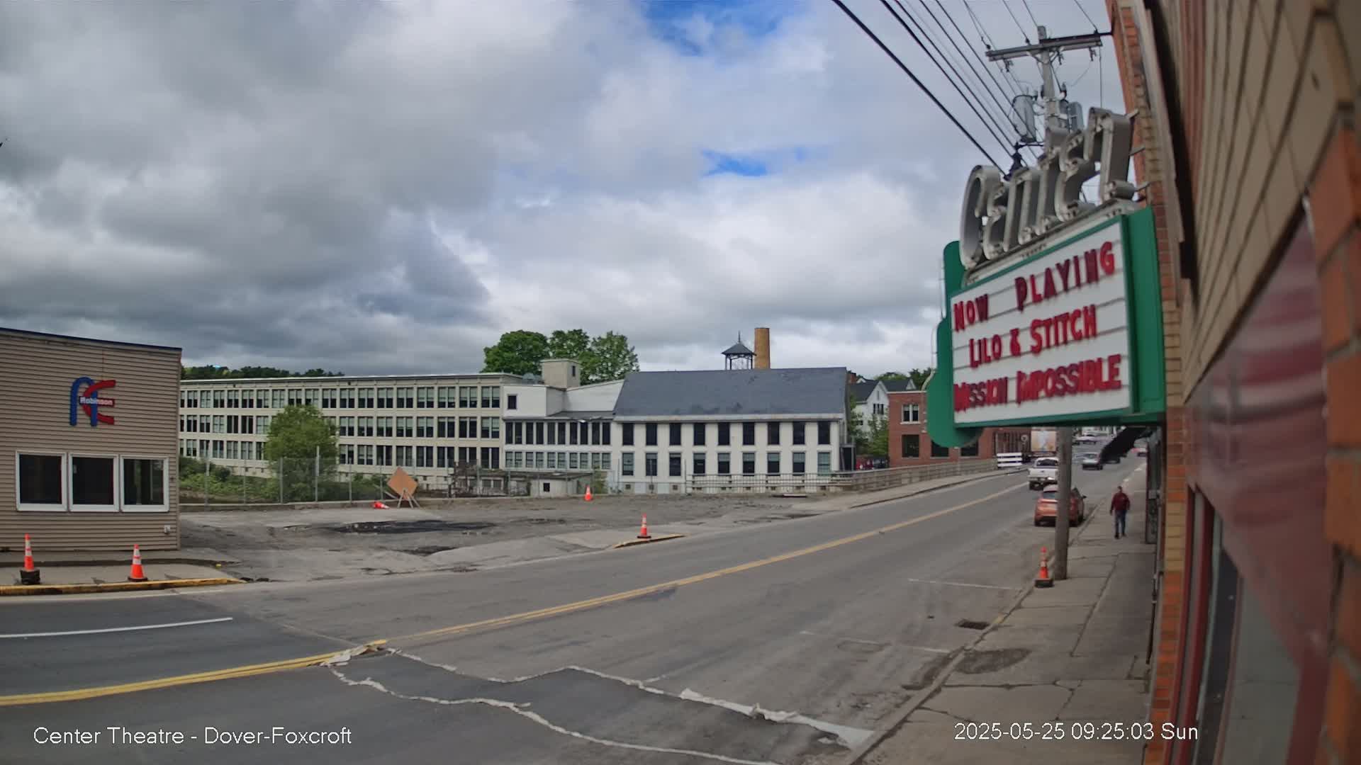 A street scene on an overcast day shows a road leading to a large industrial building across a bridge, with a movie theater marquee advertising "Lilo & Stitch" and "Mission Impossible" in the foreground.