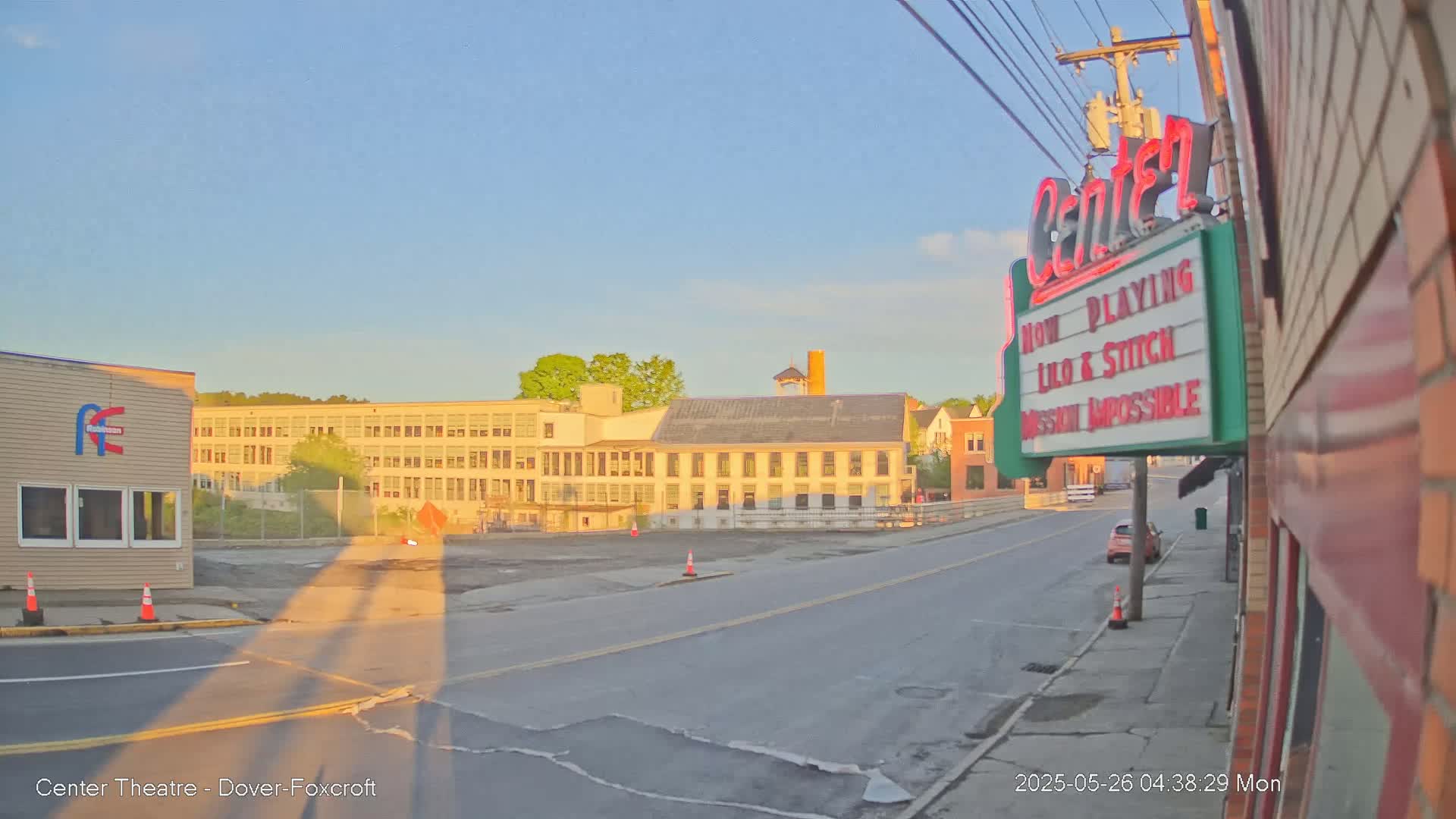 A street scene at sunrise shows a mostly empty road in front of several buildings, including a large multi-story building and a movie theater marquee.