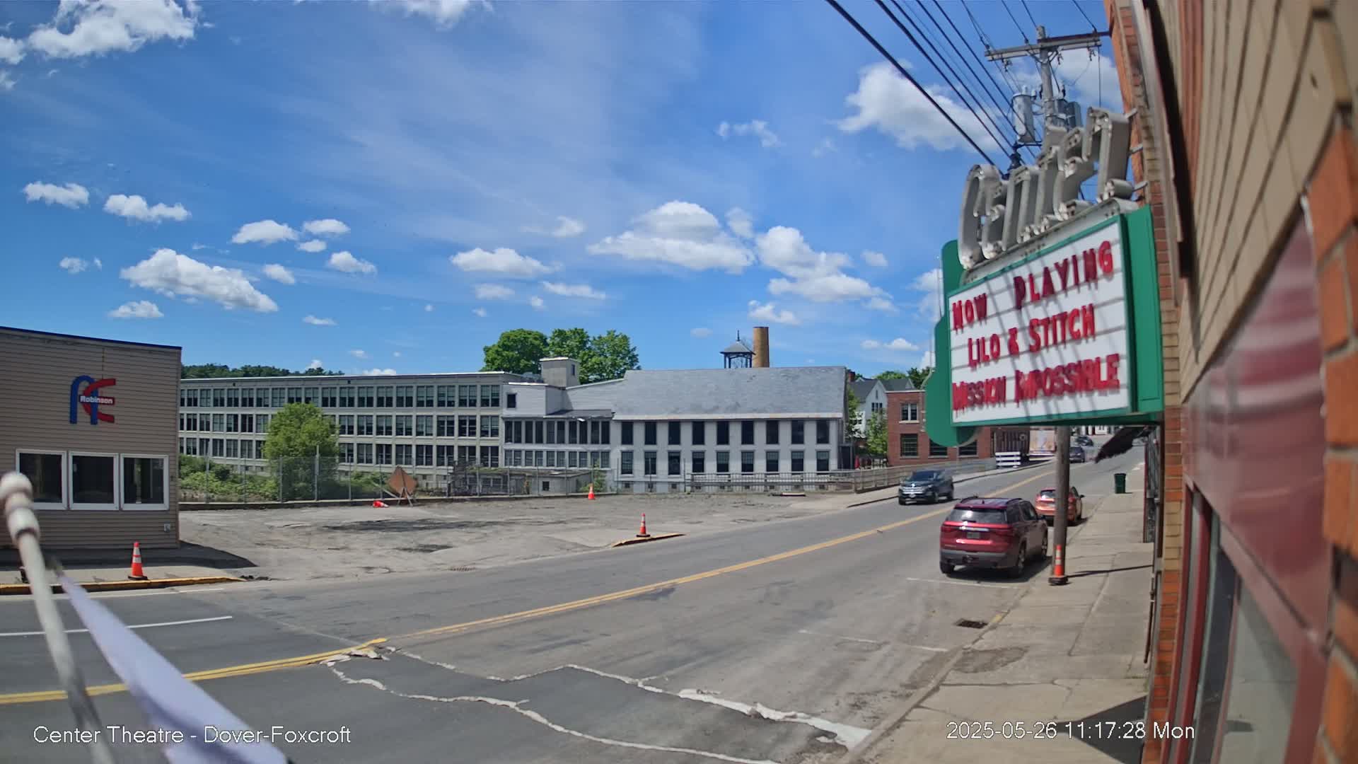 A partly sunny day shows a street scene with a large industrial building across the street from a movie theater marquee, a few cars parked and driving by.