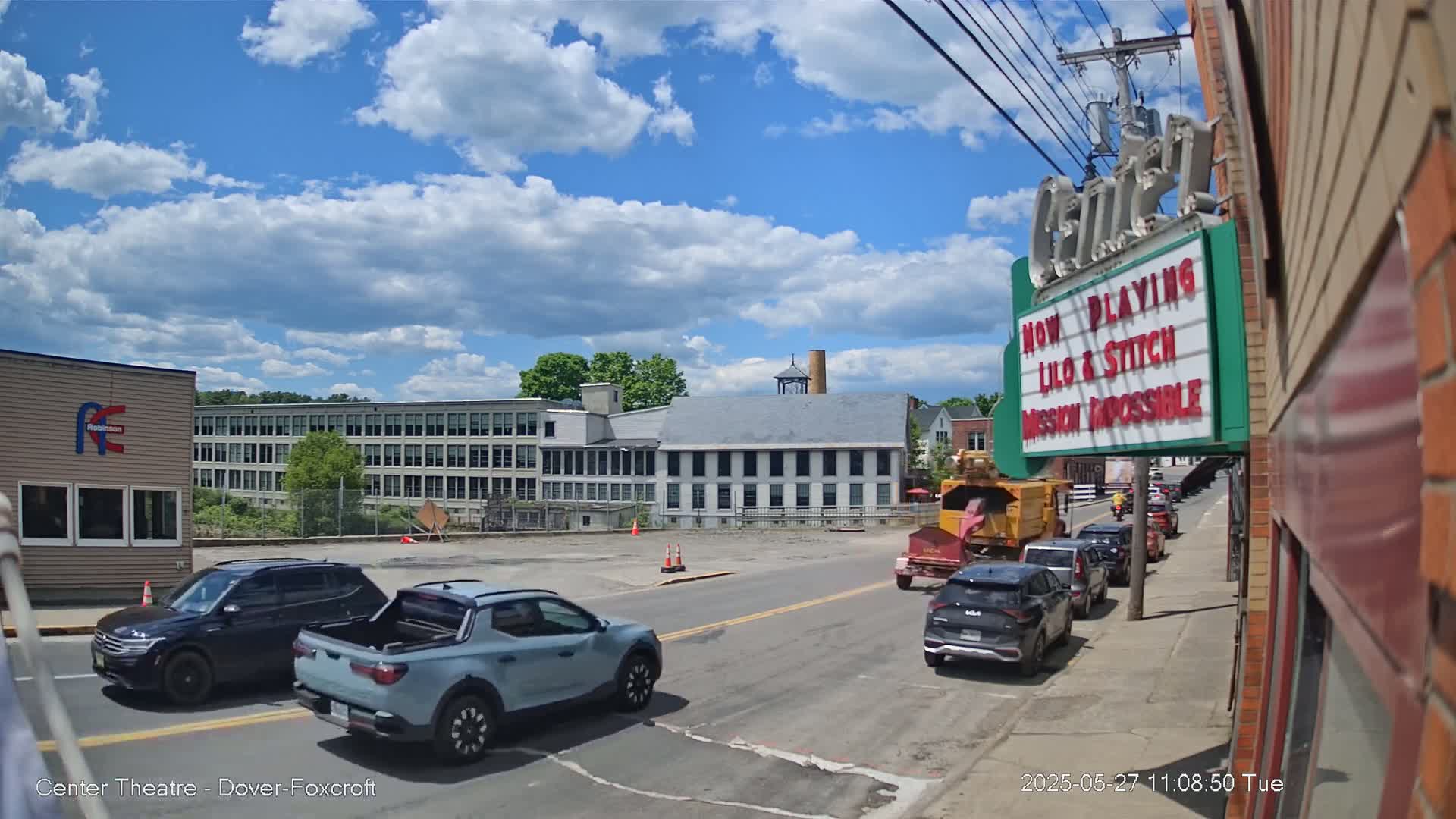 A partly cloudy day shows a street scene with several vehicles, including a light blue pickup truck, a dark SUV, and other cars parked along the side of the road, in front of a multi-story brick building and a movie theater marquee.
