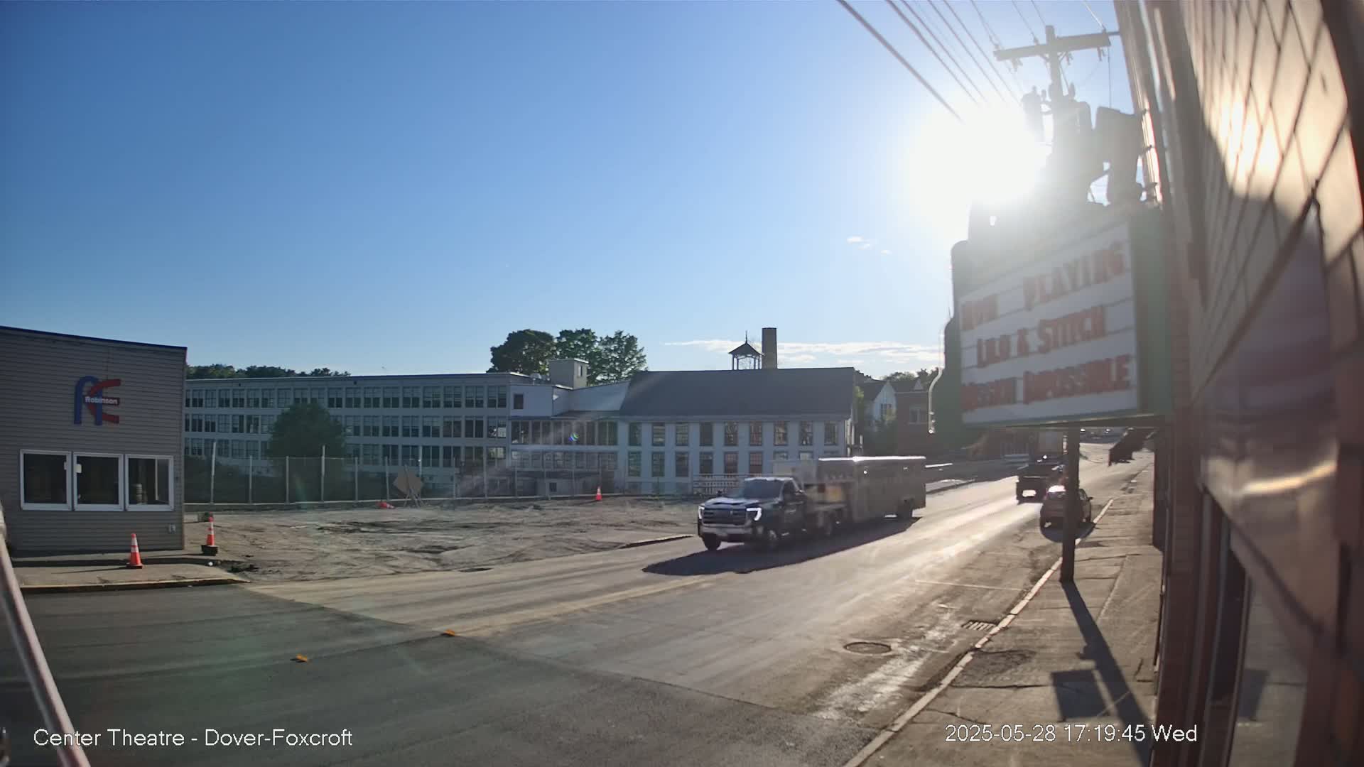 A sunny day reveals a street scene with a pickup truck towing a trailer, driving past a multi-story building under construction and other buildings, all under a clear blue sky.