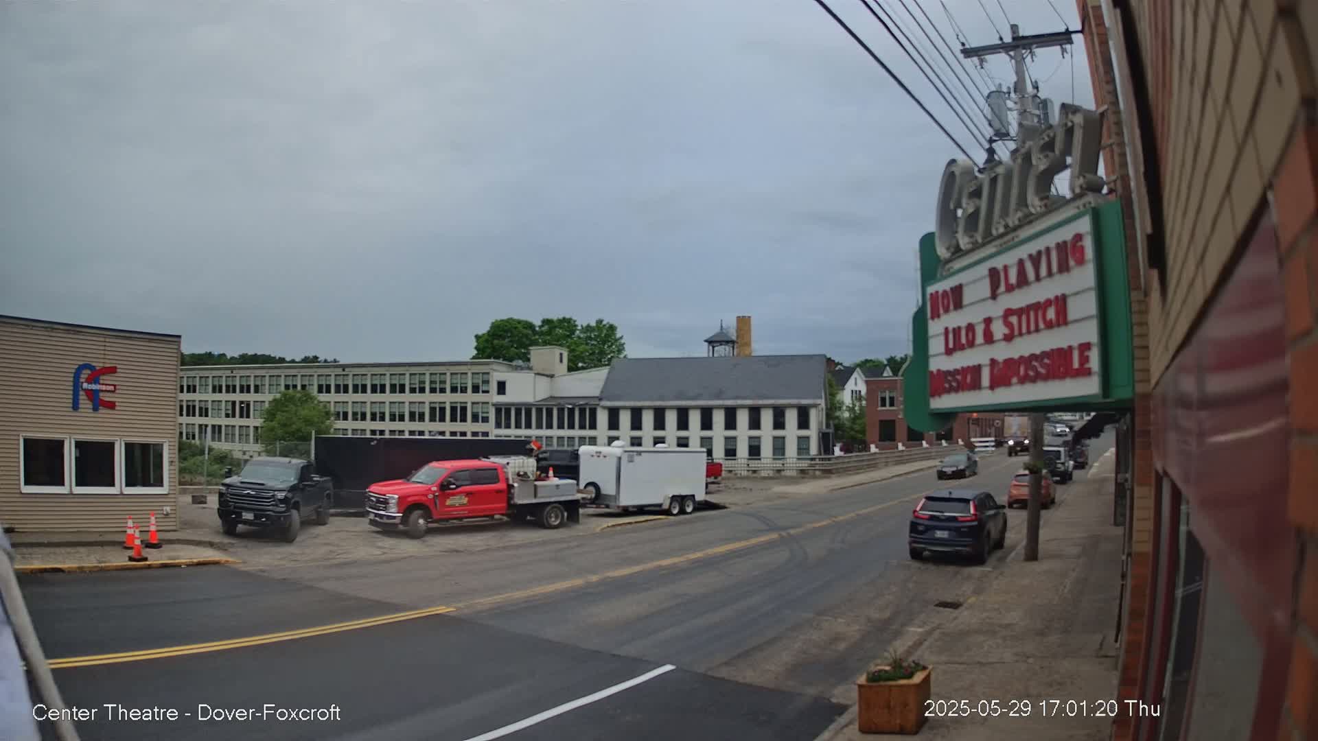 A street scene on an overcast day shows several vehicles parked near a multi-story building and a movie theater marquee advertising "Lilo & Stitch" and "Mission Impossible."
